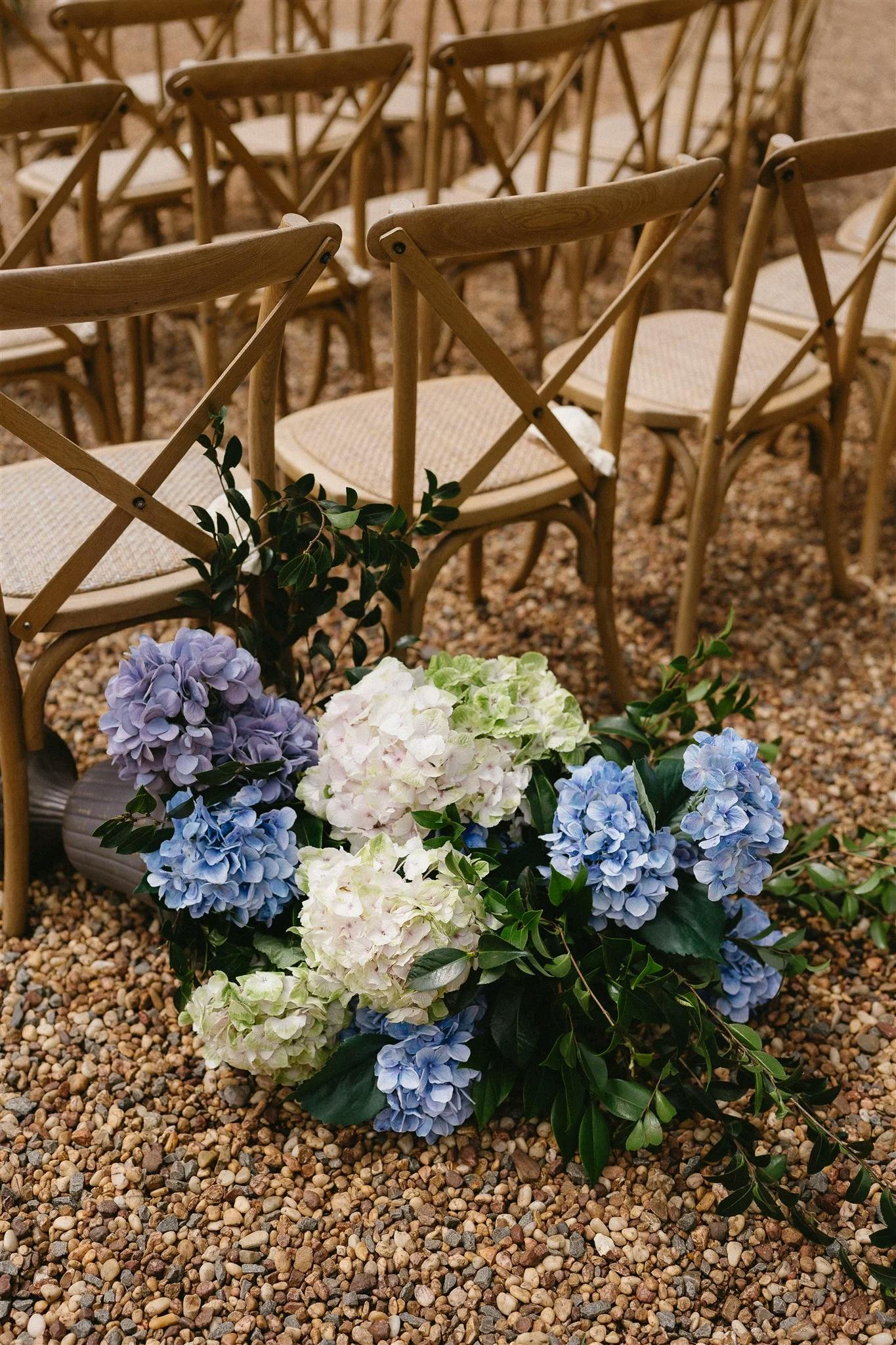 A bouquet of blue, white, and lavender hydrangeas with green foliage lies on a gravel pathway next to rows of wooden chairs with woven seats, likely arranged for an outdoor event or ceremony.