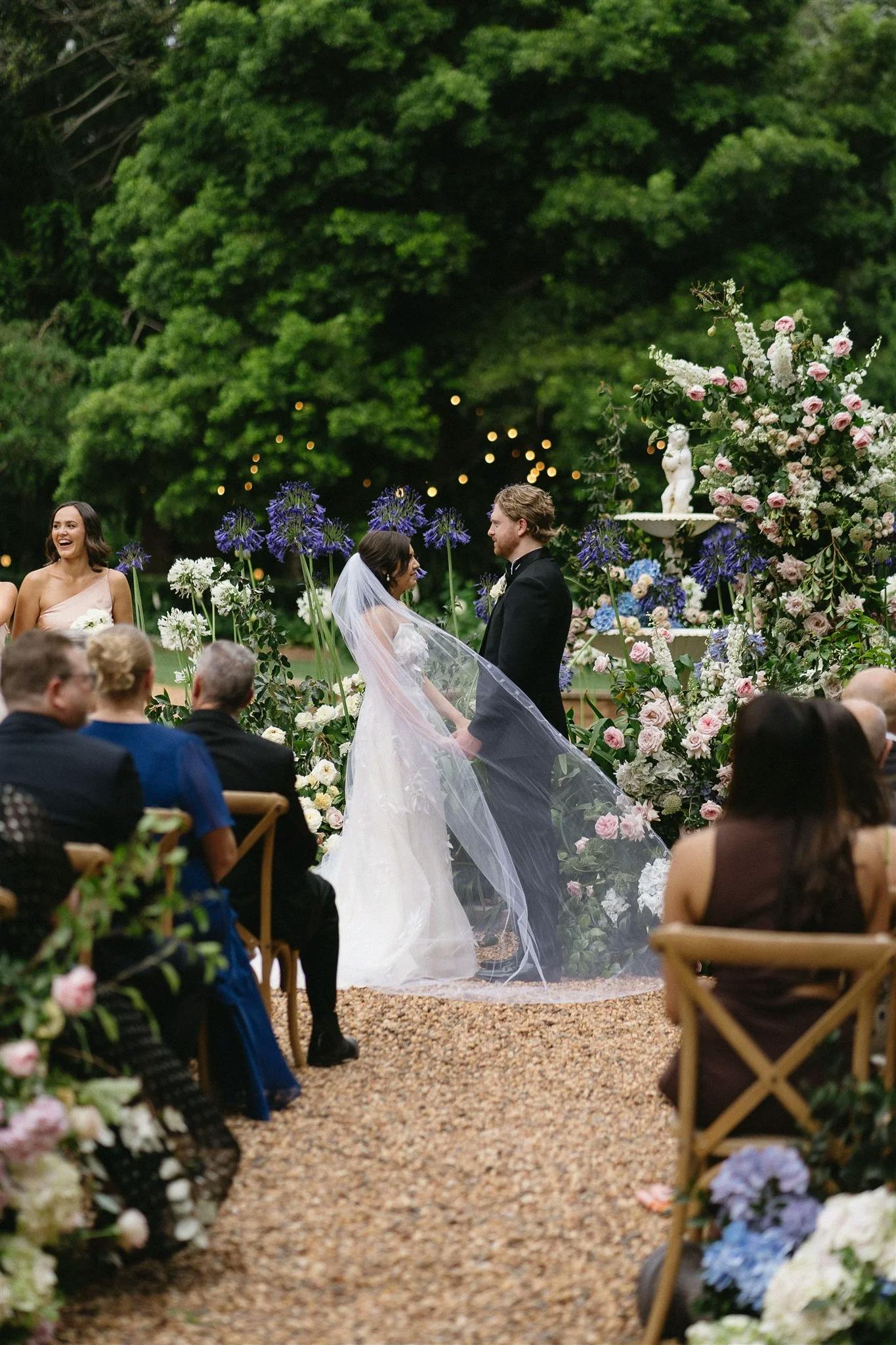 A couple stands at the altar during an outdoor wedding ceremony, surrounded by lush greenery and floral arrangements. The bride and groom face each other, holding hands. Guests are seated on either side, attentively watching the ceremony.