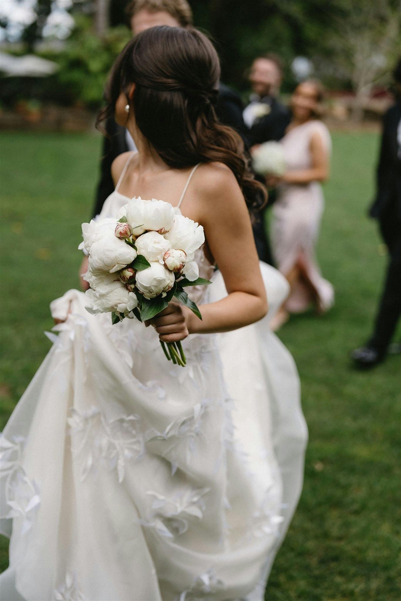 A bride holding a bouquet of white flowers walks on a grassy lawn. She is seen from behind, wearing a white dress with floral details. In the blurred background, a man in a suit and two women, one in a light-colored dress, are also visible.