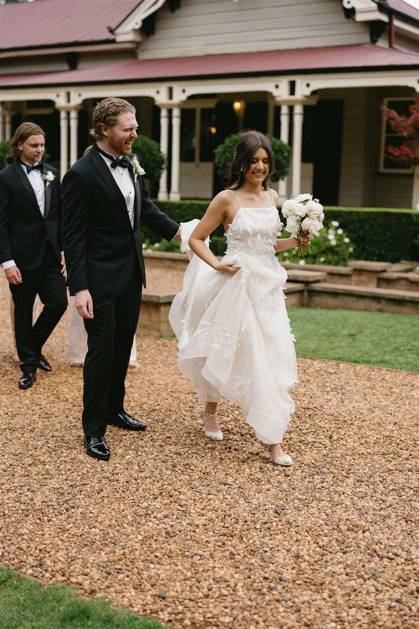 A bride in a white dress holds a bouquet and is followed by a groom in a black tuxedo and a groomsman as they walk on a gravel path in front of a charming house with a maroon roof and white porch.