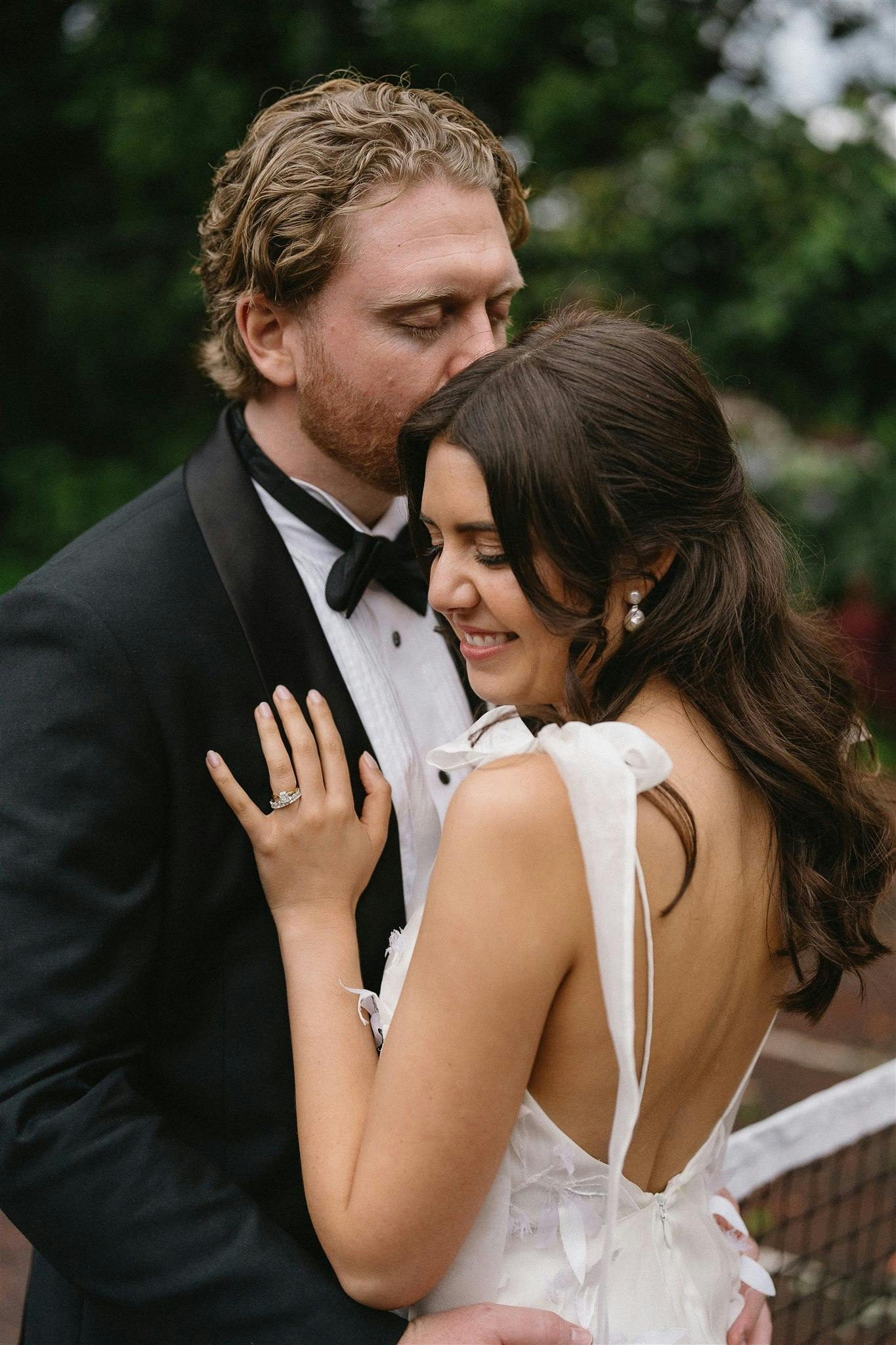 A groom in a black tuxedo tenderly embraces his smiling bride, who is wearing a white dress with a deep back and ribbon details. They stand outdoors in a lush setting, with greenery visible in the background. The groom's eyes are closed as he holds his bride close.