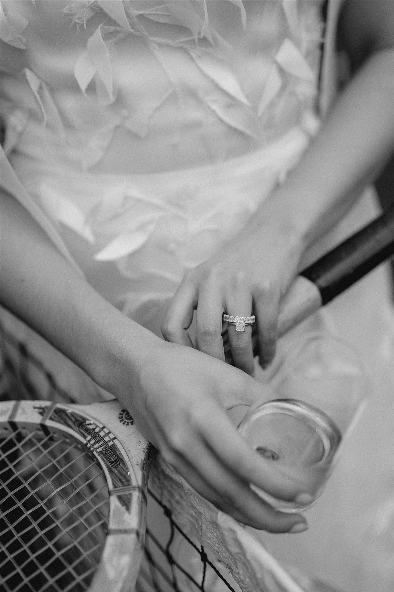 Black and white photo of a person holding a wine glass in one hand and resting the other hand on a tennis racket. They are wearing a dress with floral accents and a ring on their left ring finger.