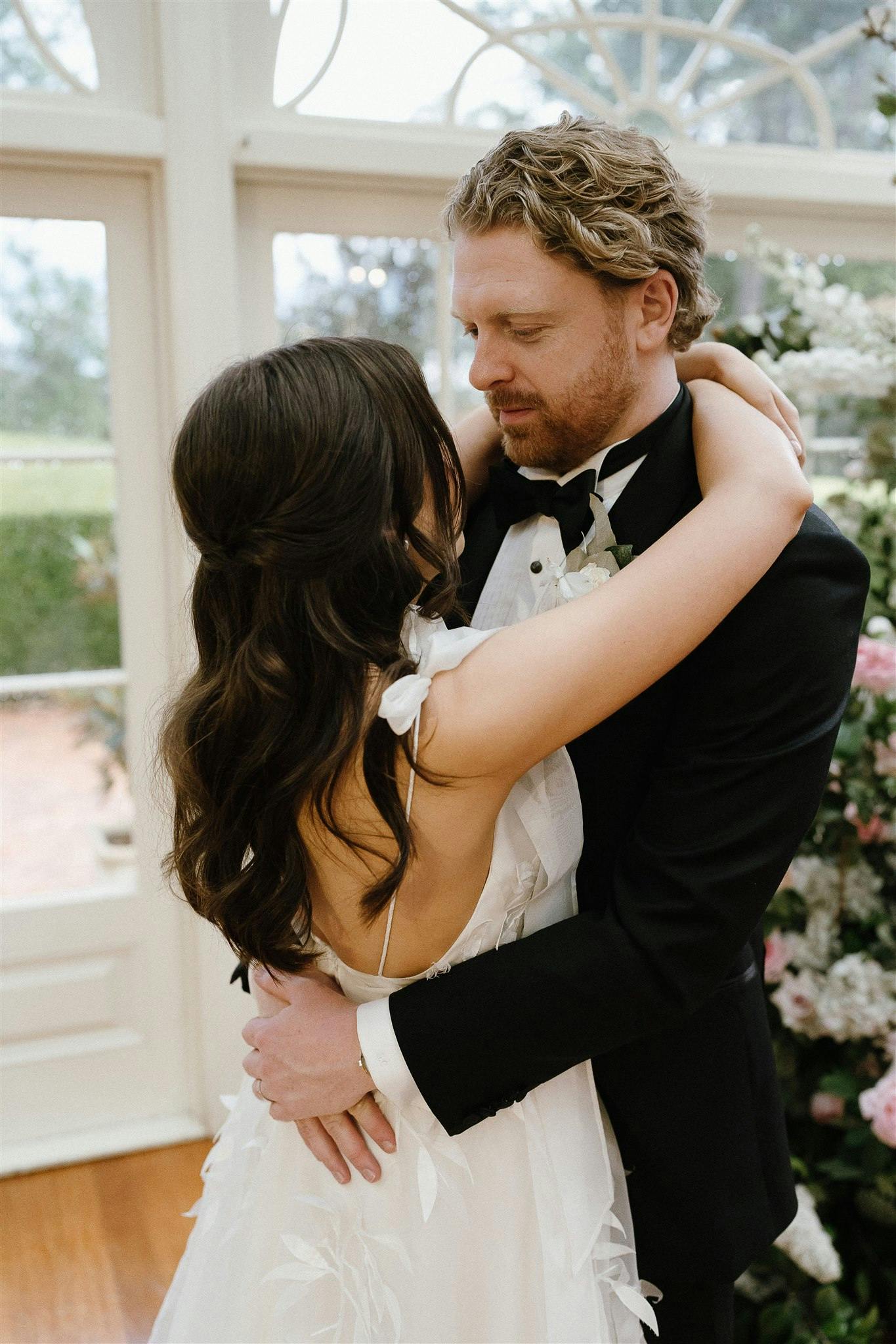 A couple dressed in wedding attire embrace each other lovingly in front of a window. The bride is wearing a white dress with her back to the camera, while the groom, in a black suit, gazes down at her. Flower decorations fill the background, creating a romantic scene.