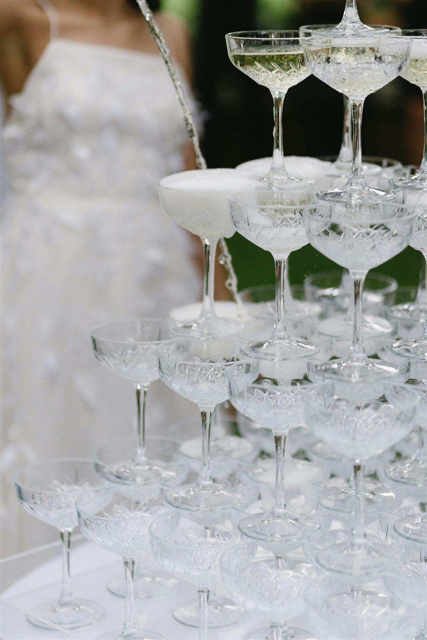 A close-up of a champagne tower consisting of multiple layers of stacked crystal glasses being filled from the top. The cascading champagne creates a foamy overflow. A person in a white dress stands in the blurred background.