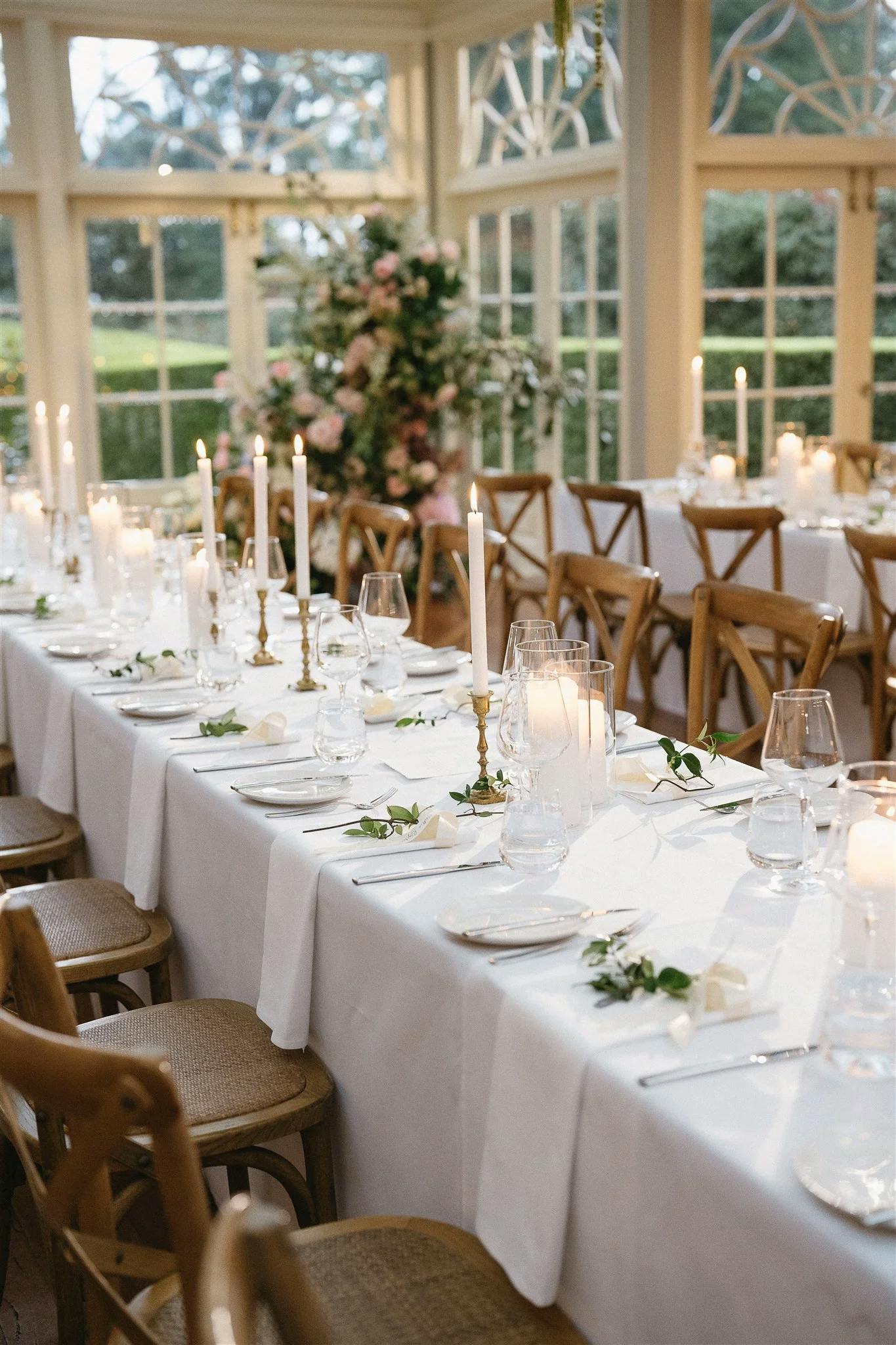 A beautifully decorated dining table with white tablecloth and elegant place settings, featuring tall white candles, clear glassware, and greenery accents. The table is set in a bright conservatory with large windows and ornate garden views in the background.