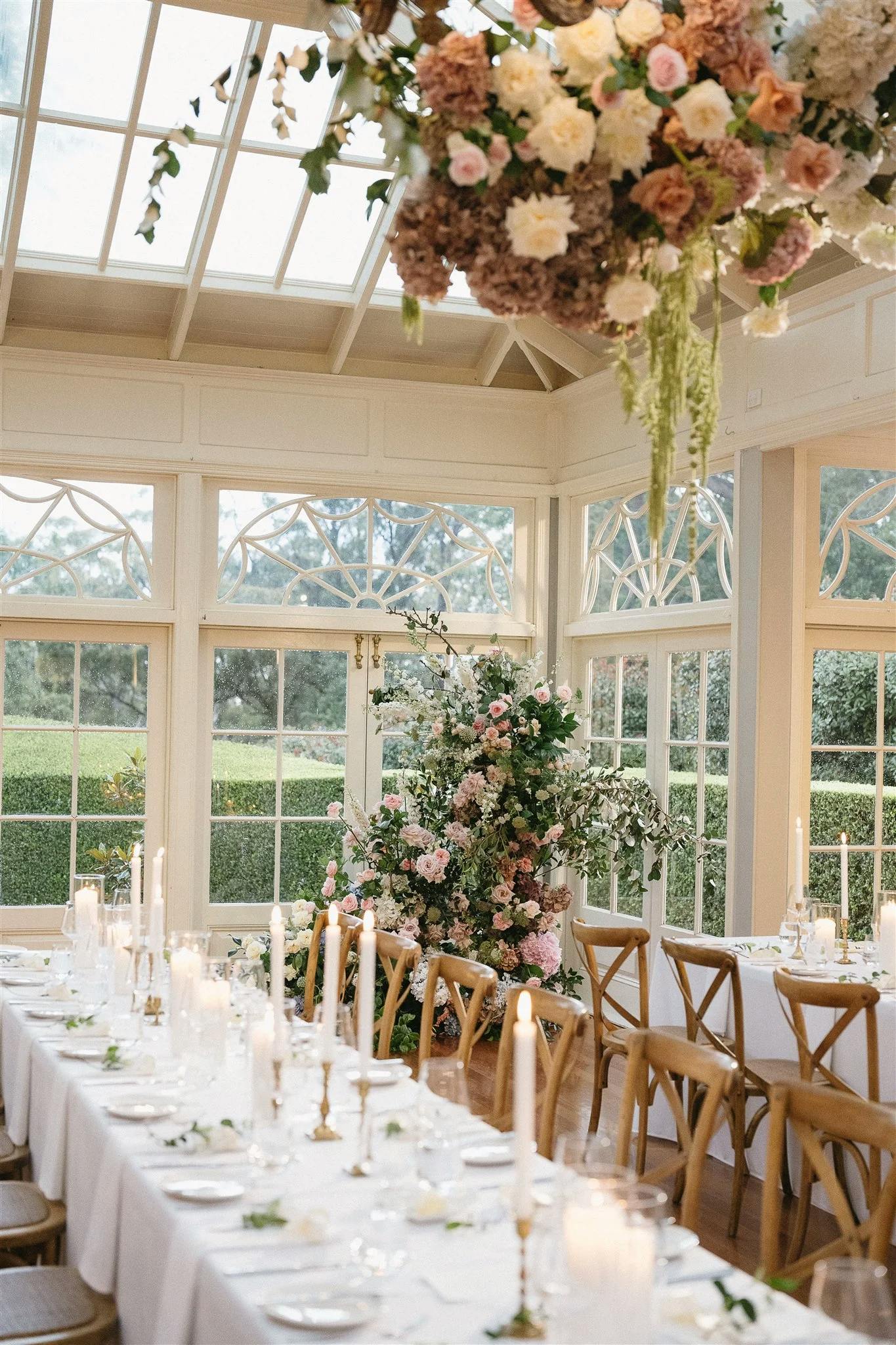 A beautifully decorated wedding reception room with floor-to-ceiling windows. Long tables are adorned with white cloths, numerous lit white candles, and elegant floral arrangements. A large hanging floral arrangement is above the table, adding to the ambiance.