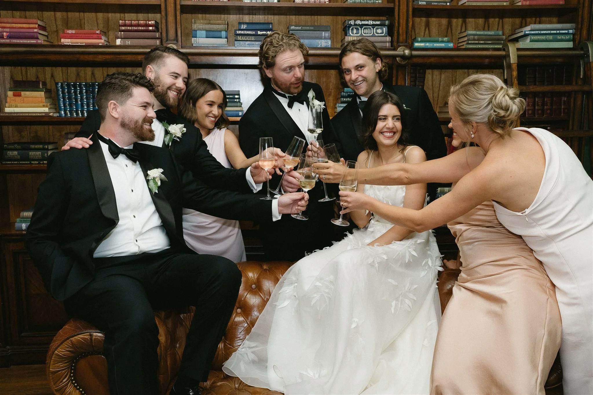 A group of seven people dressed in formal attire are sitting and standing around a leather couch, raising their glasses in a celebratory toast. They are all smiling and appear to be enjoying the festivity in a room lined with bookshelves.