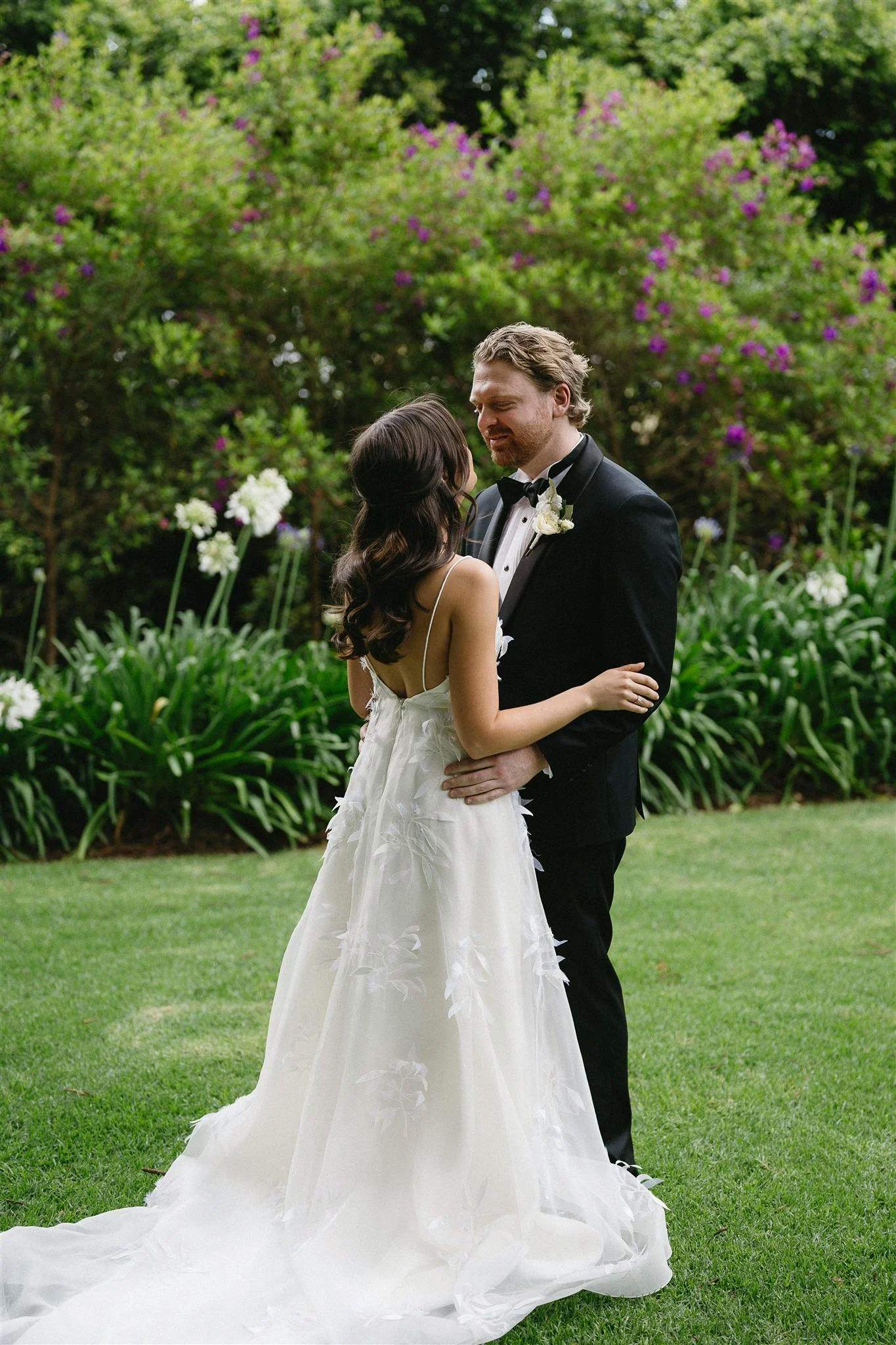 A bride and groom share an intimate moment outdoors. The bride wears a white gown with a long train, and the groom is dressed in a black tuxedo with a white bow tie. They are standing on a grassy lawn, surrounded by greenery and blooming flowers.