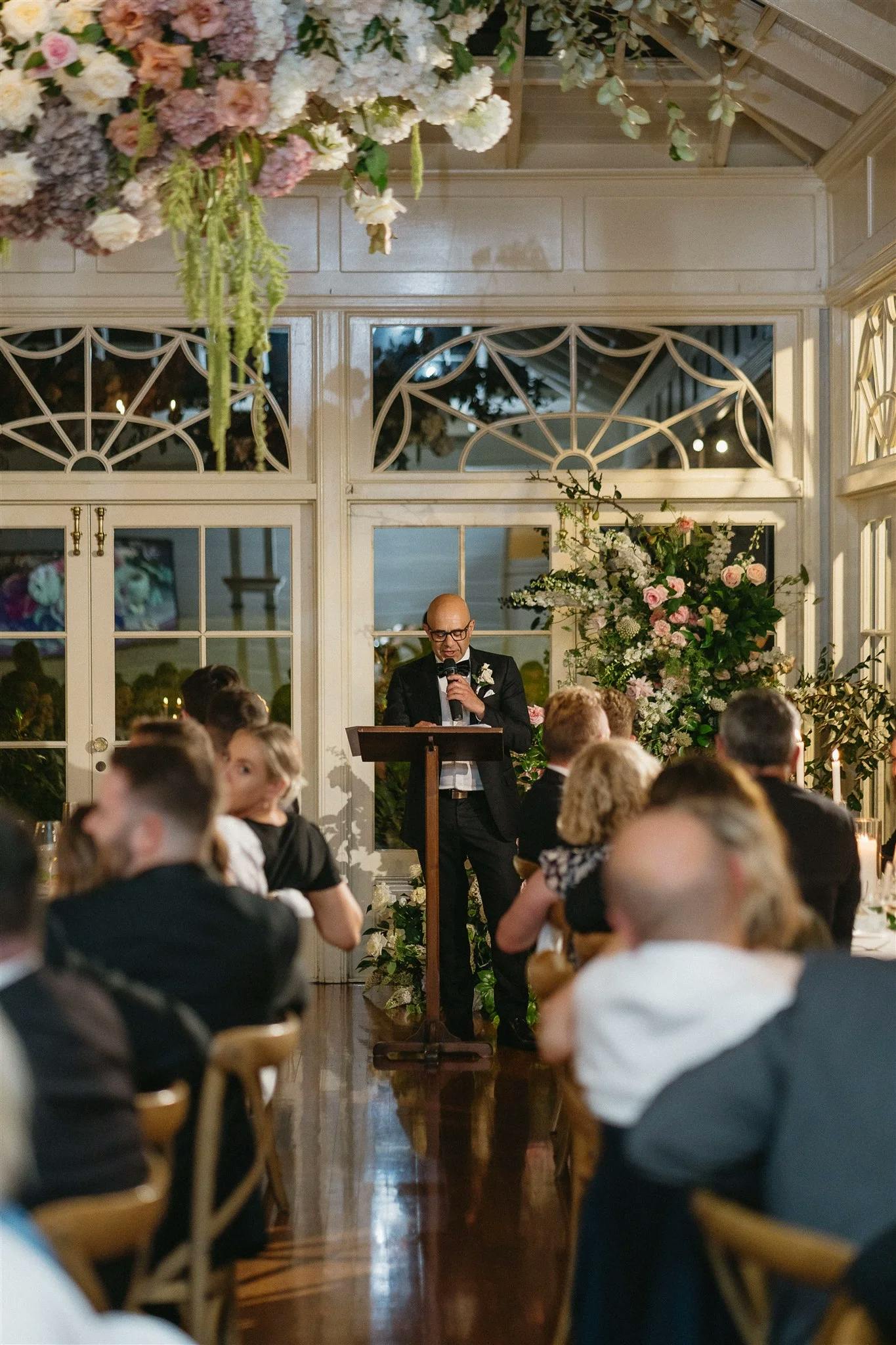 A person standing at a lectern speaks to an audience seated at tables in a well-decorated room with large windows and abundant floral arrangements hanging from the ceiling and placed around the area. The setting appears elegant and formal.