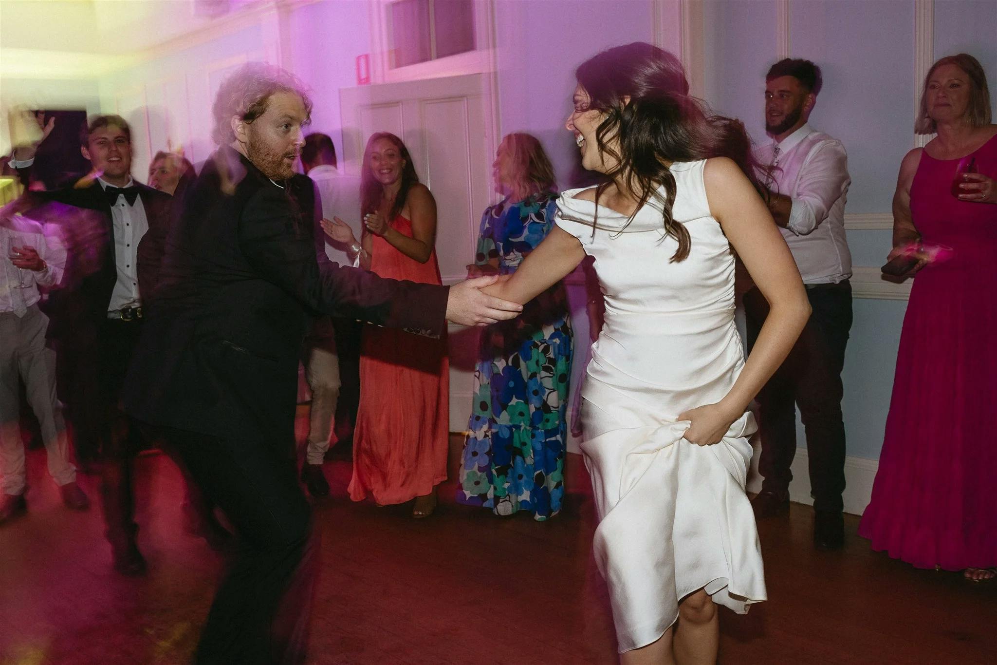 A man and woman dance energetically at a party, with the woman in a white dress and the man in a suit. Around them, other guests in colorful outfits are talking and watching. The lighting is soft and colorful, creating a lively atmosphere.