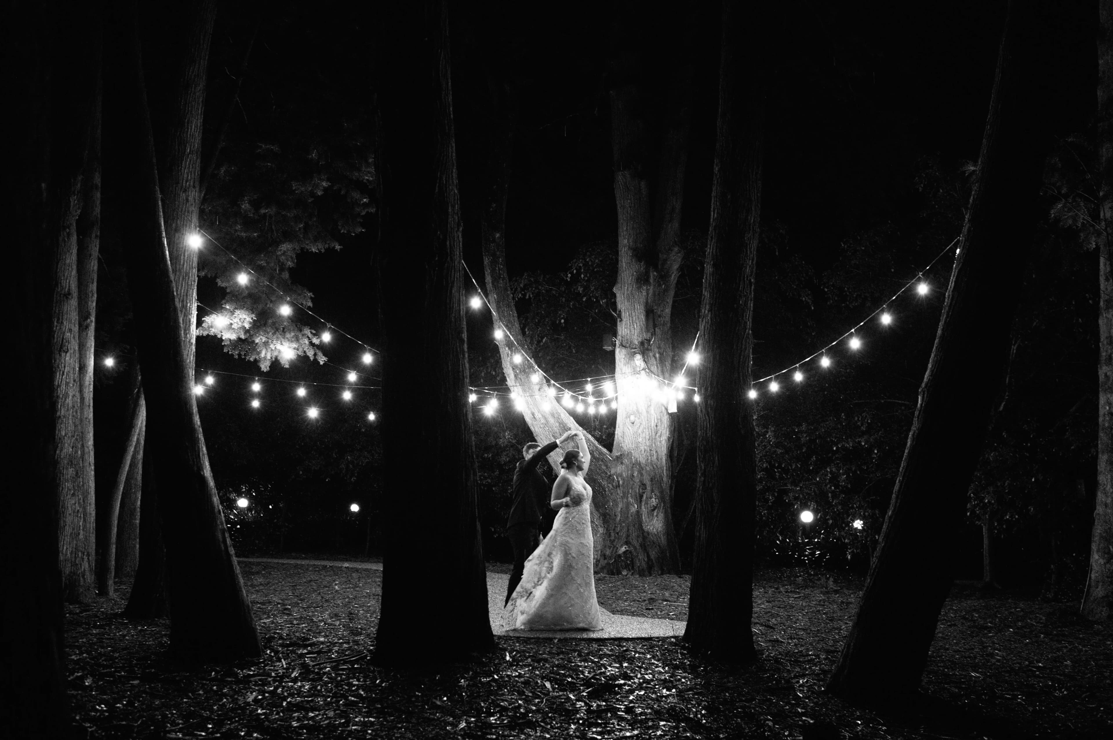 A bride and groom stand under a canopy of string lights in a wooded area at night. The scene is illuminated by the soft glow of the lights, creating a romantic atmosphere. The background is dark, emphasizing the couple and the lights.