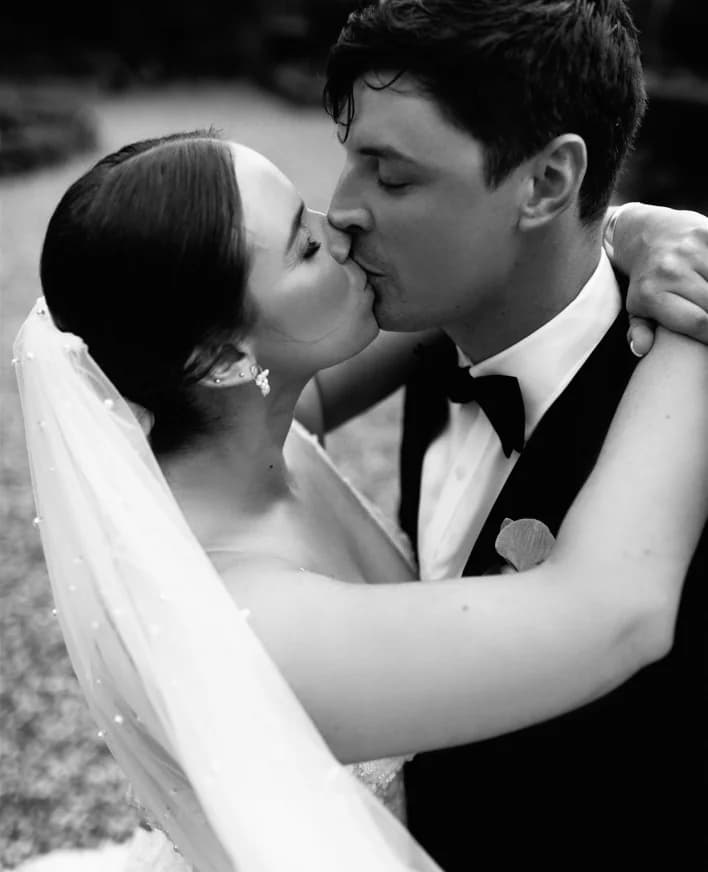 A bride and groom share a kiss on their wedding day. The bride wears a veil and earrings, while the groom is in a tuxedo with a bow tie. They embrace lovingly against a blurred outdoor background.