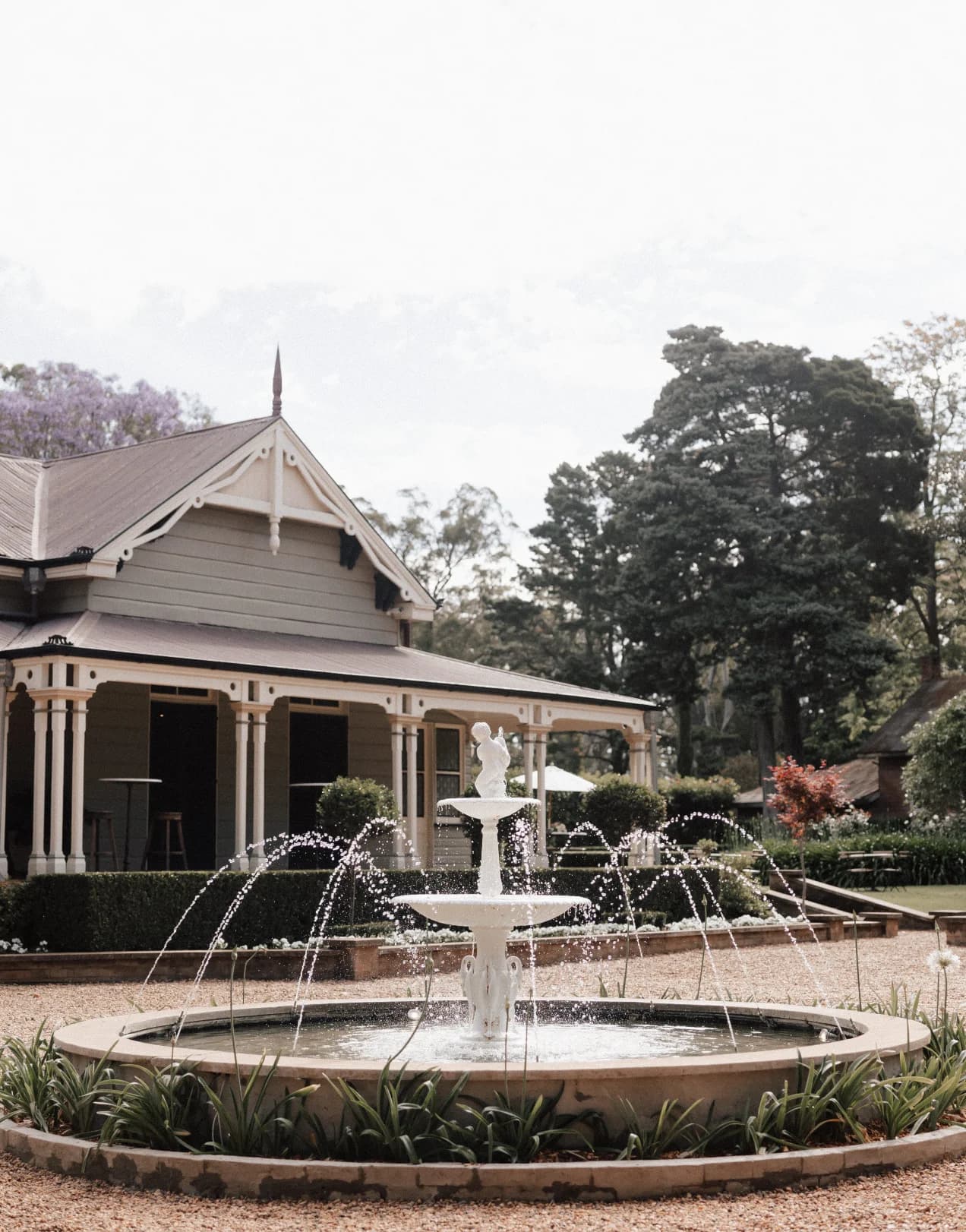 A charming, rustic house with a wraparound porch is surrounded by trees and a garden. In the foreground, a tiered white fountain gracefully sprays water, adding to the serene ambiance of the scene.