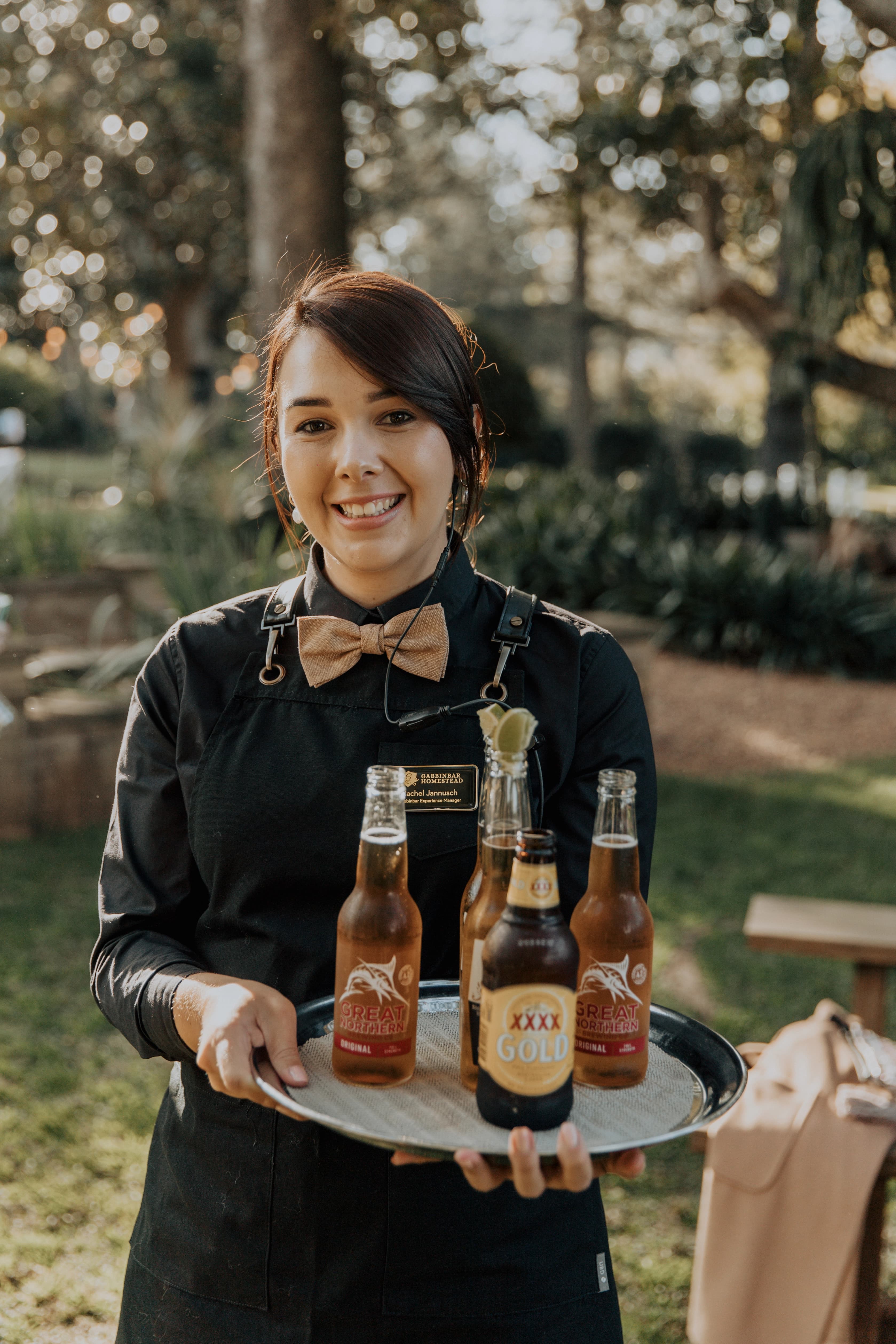 A server in a black uniform and bow tie is holding a tray with three beer bottles. She is smiling and standing outdoors, with trees and a lawn visible in the background.
