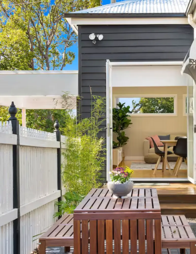 A cozy outdoor patio features a wooden table with a potted plant, adjacent to a white picket fence. A door leads into a light-filled interior with plants and a wooden dining table, surrounded by vibrant greenery under a clear blue sky.