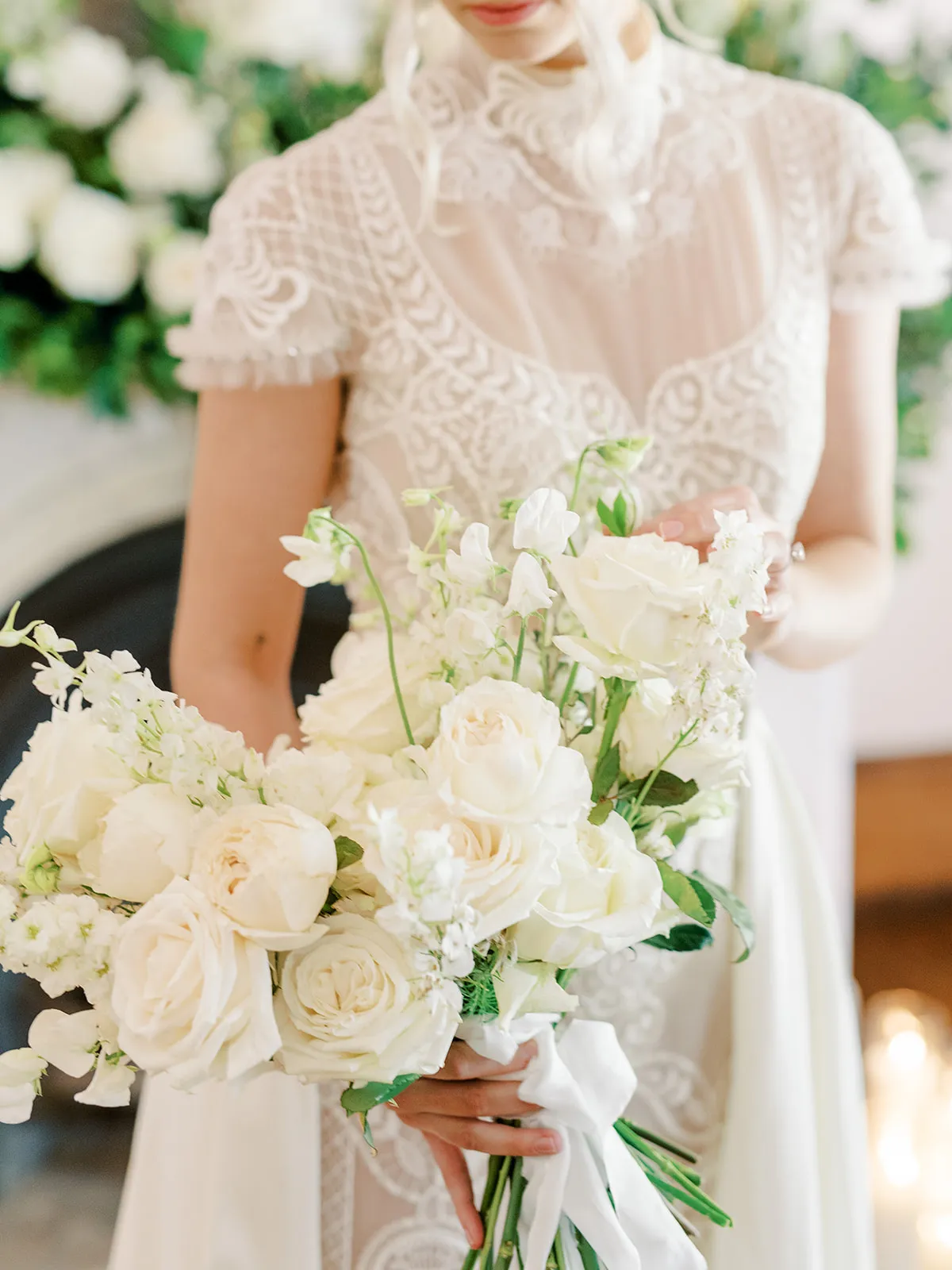 A bride in a lace wedding dress holds a bouquet of white flowers. The detailed lace pattern of her dress is visible, and the bouquet includes various white blooms, some with greenery. The background features soft-focus floral decorations.