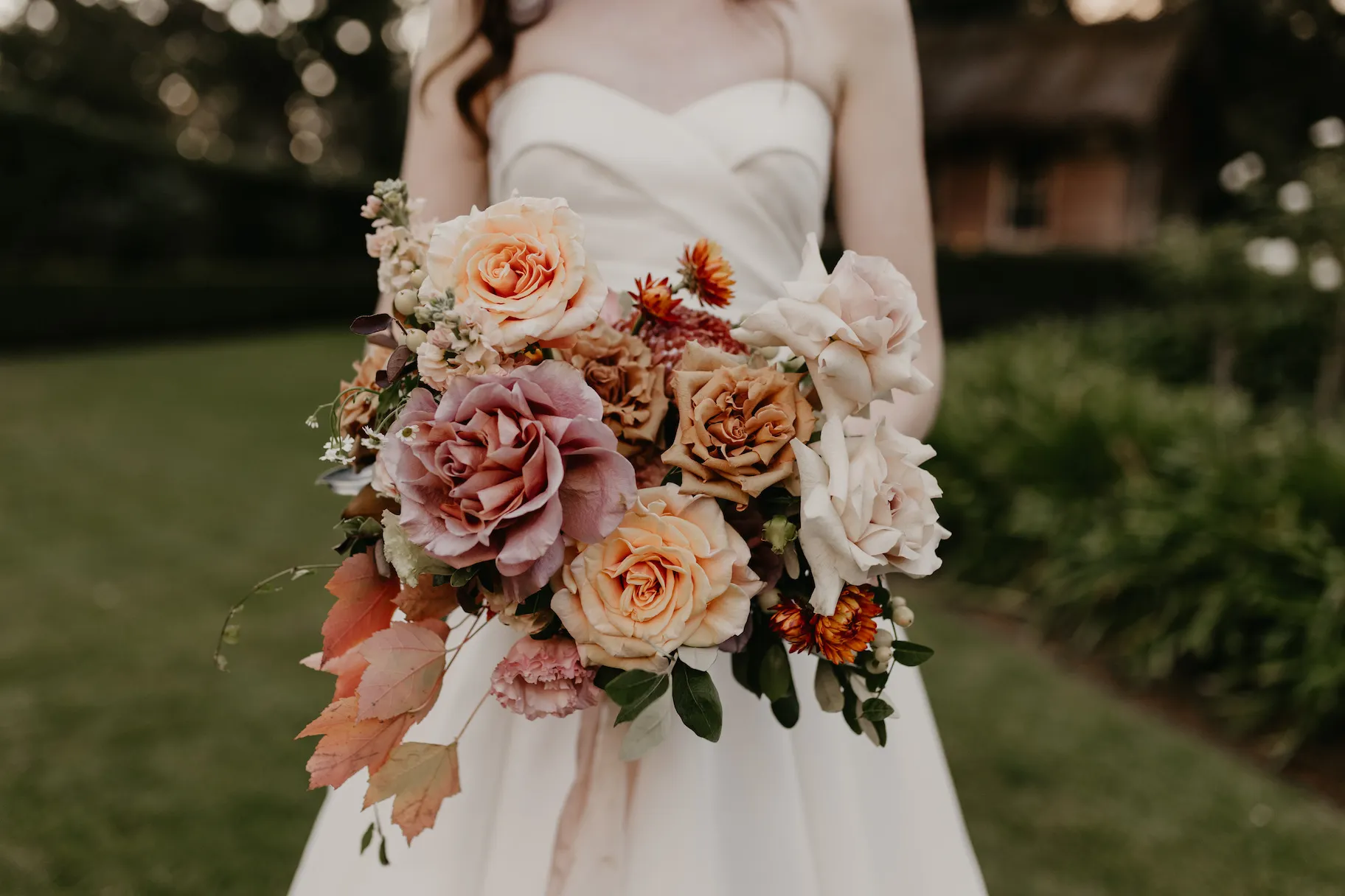 A bride in a strapless white dress holds a large, colorful bouquet featuring a mix of pastel roses, orange flowers, and greenery. The background shows a garden setting with green grass and an out-of-focus rustic building.