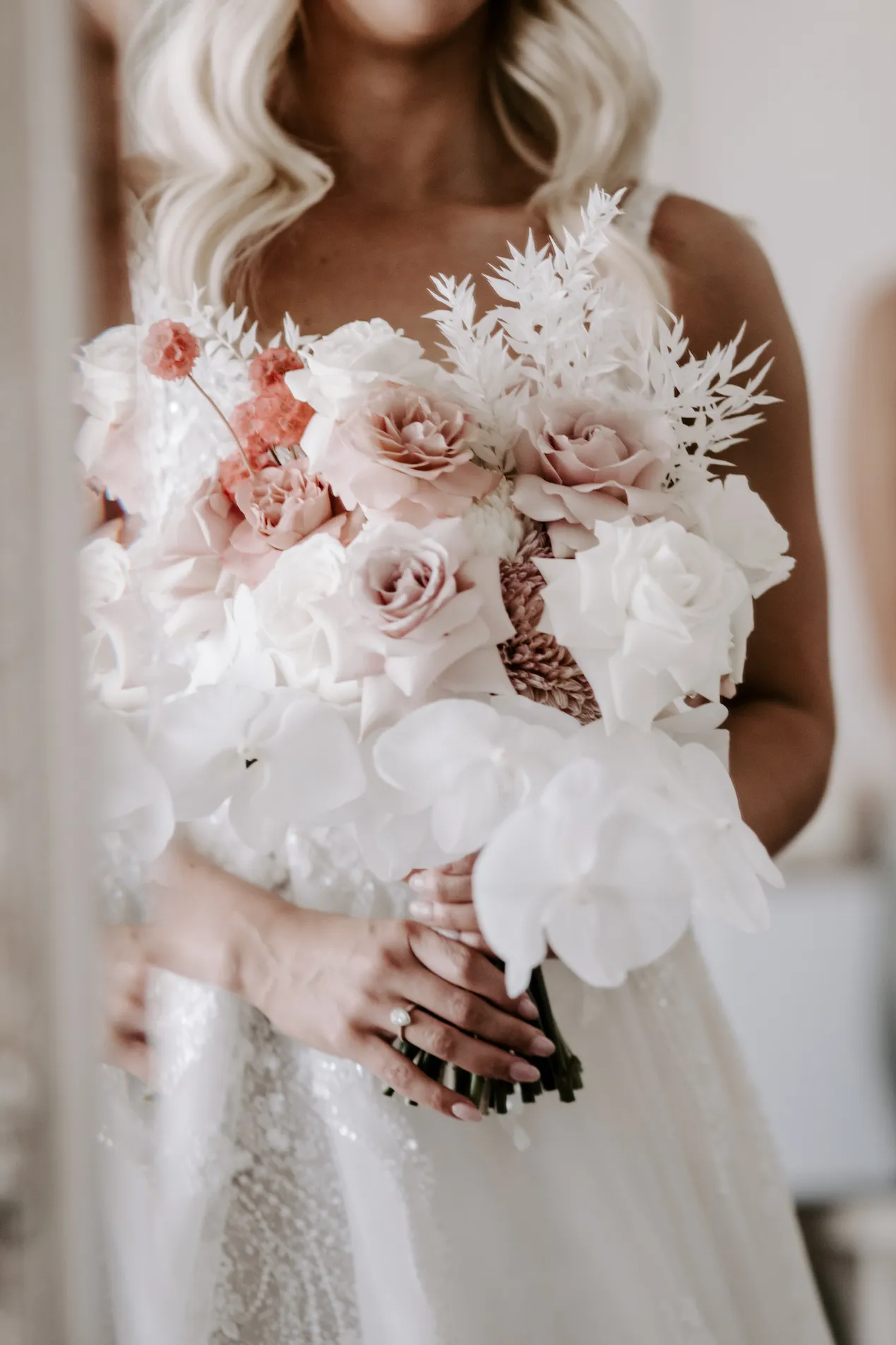 A bride in a wedding dress holds a floral bouquet featuring white orchids, pink roses, and other assorted flowers and greenery. The focus is on the flowers, with the bride's face partially out of frame. The image has a soft, romantic feel.