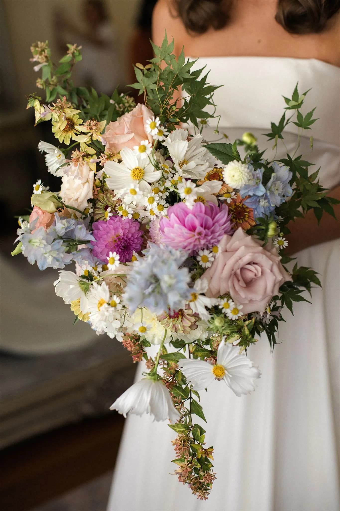 A bride holding a colorful bouquet featuring an assortment of flowers, including roses, cosmos, dahlias, and other blooms, along with green foliage. She is wearing a strapless white wedding dress.