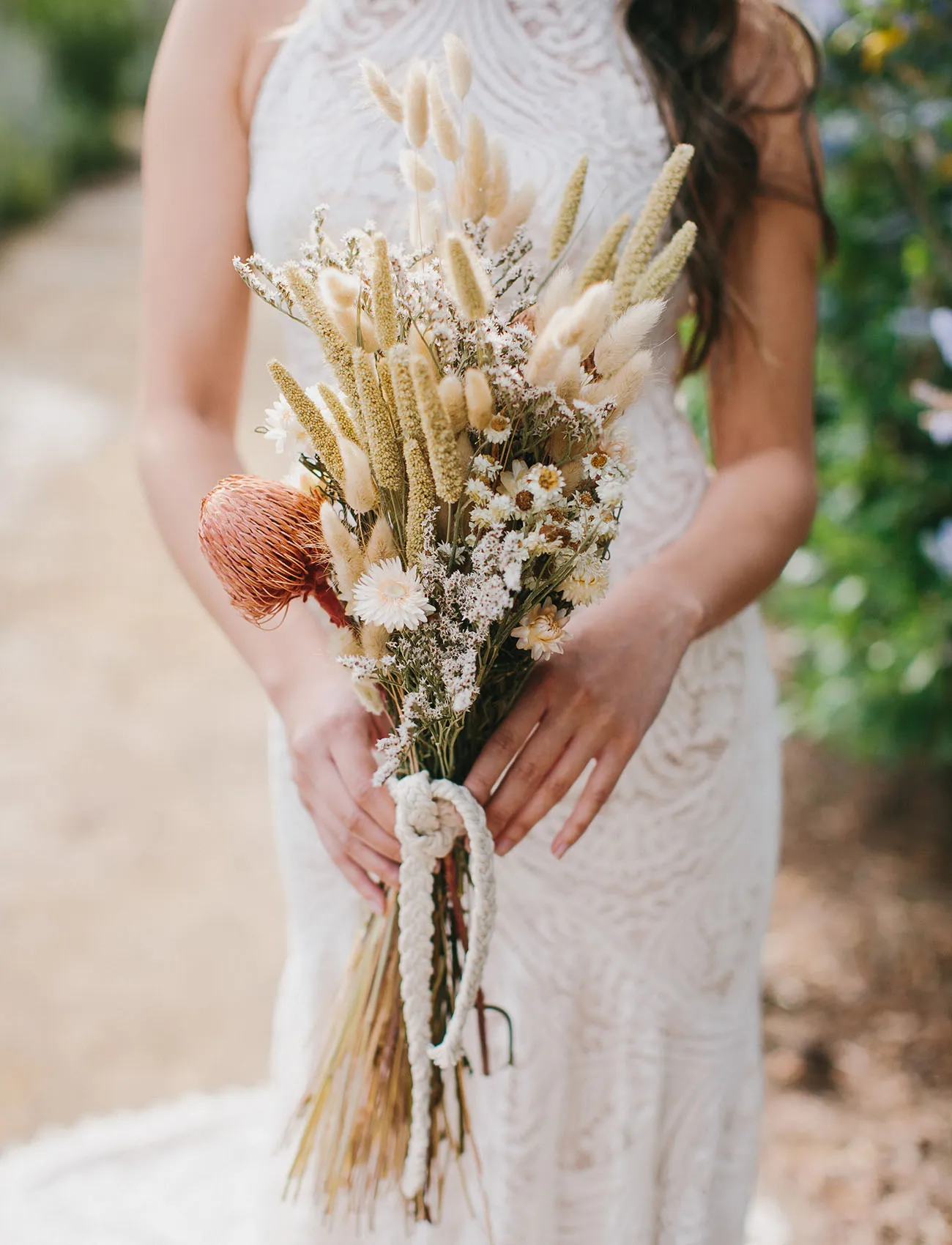 A person in a white lace dress is holding a bouquet of dried flowers, including beige and white blooms, and a single red flower. The bouquet is tied with a white rope, and greenery can be seen in the background.