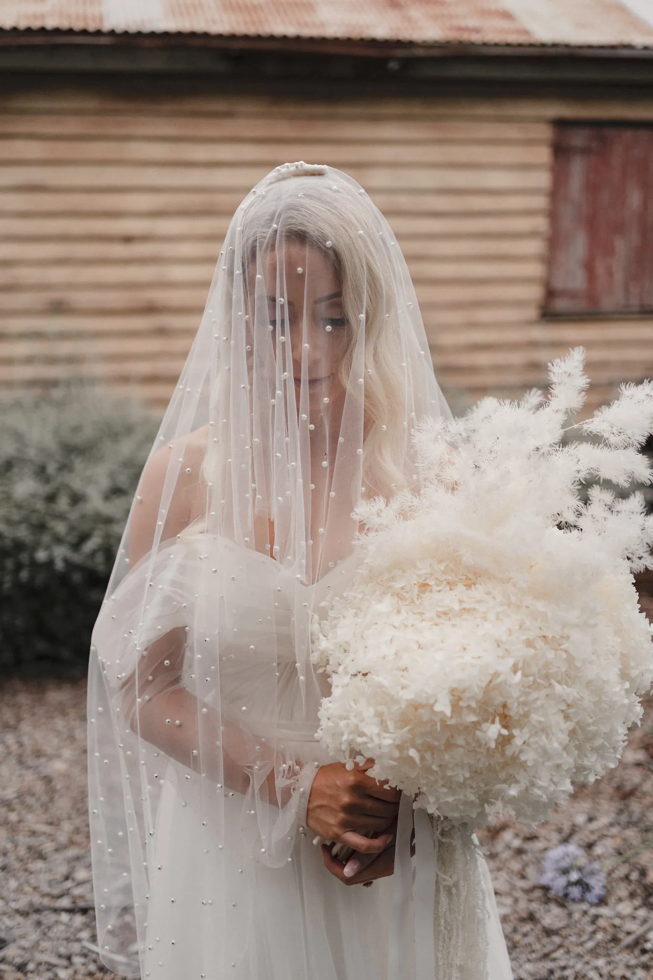 A bride dressed in a white wedding gown and a veil adorned with small pearls holds a large bouquet of white flowers. She stands outdoors in front of a rustic wooden building, looking down thoughtfully.