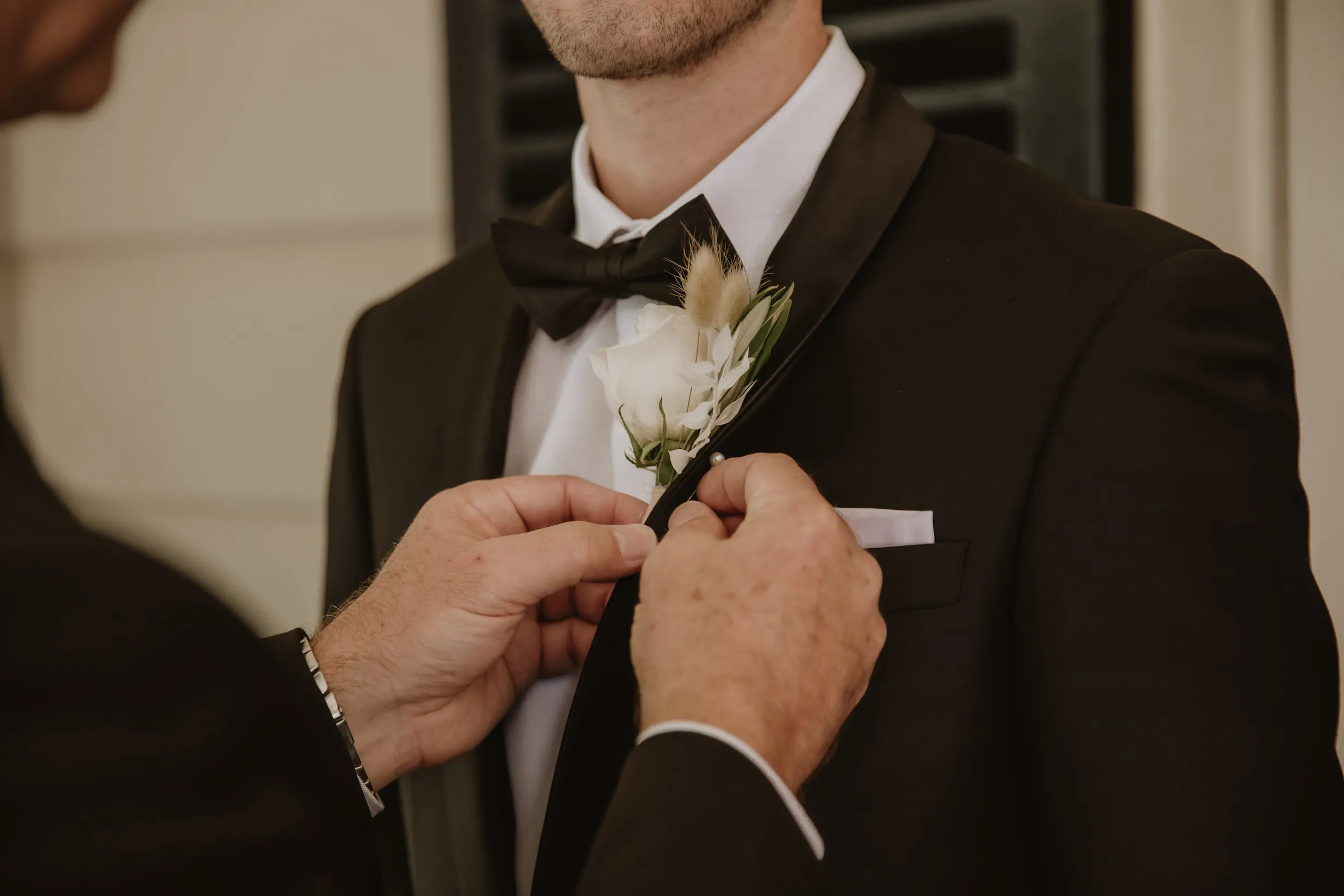 A person wearing a black tuxedo and black bow tie is having a white flower boutonniere pinned to their lapel by another individual. The background is softly blurred, emphasizing the focus on the attire and the action.