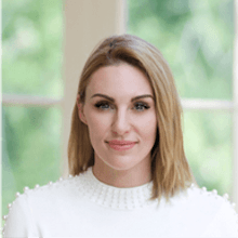A woman with shoulder-length blonde hair and a white sweater with pearl embellishments is smiling subtly at the camera. The background is blurred, showcasing a green, leafy outdoor scene seen through large windows.