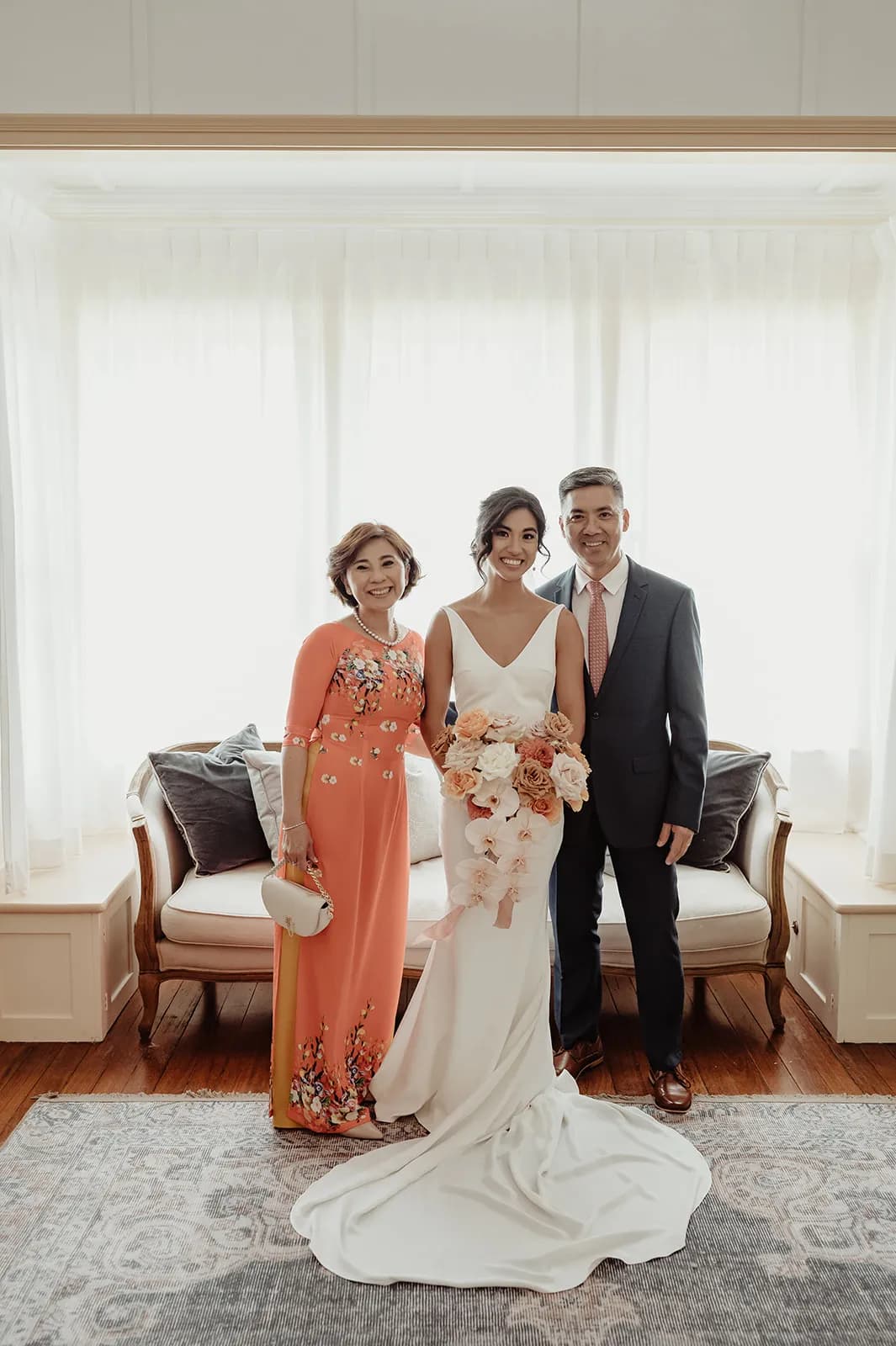 A bride stands between two older adults in a well-lit room. She wears a white wedding dress and holds a bouquet of flowers. The woman on her right wears a floral-patterned red dress. The man on her left wears a dark suit with a pink tie. They are all smiling.