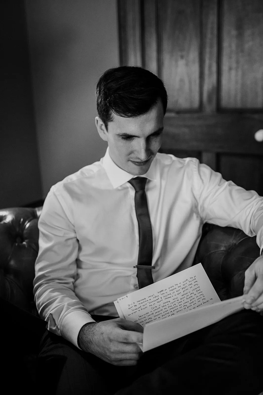 A black and white image of a man sitting on a cushioned chair, dressed in a white shirt and dark tie. He is focused on reading a handwritten letter in his hands. A wooden door is partially visible in the background.