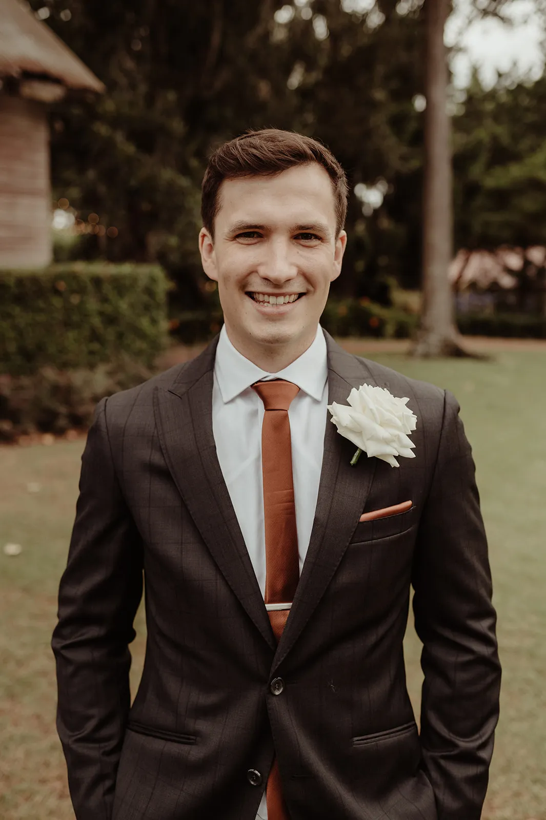 A man in a dark suit with a white shirt and a brown tie stands smiling outdoors. He has a large white rose pinned to his lapel and a brown pocket square. The background features greenery and a building with a rustic appearance.