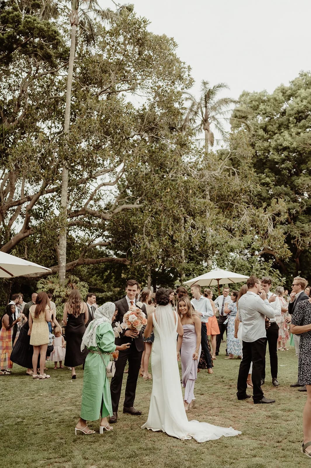 A group of people stand and mingle in a garden setting, with trees and greenery in the background. A bride in a white gown is at the center, talking to guests. Attendees are dressed in semi-formal attire, engaging in conversation and festivities under overcast skies.