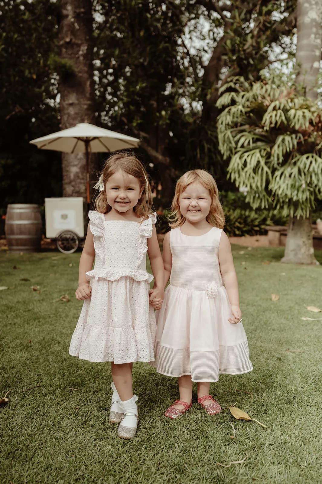 Two young girls are standing on grass and holding hands, smiling at the camera. Both are wearing light pink dresses and have light brown hair. In the background, there are trees, a large plant, and a white umbrella.