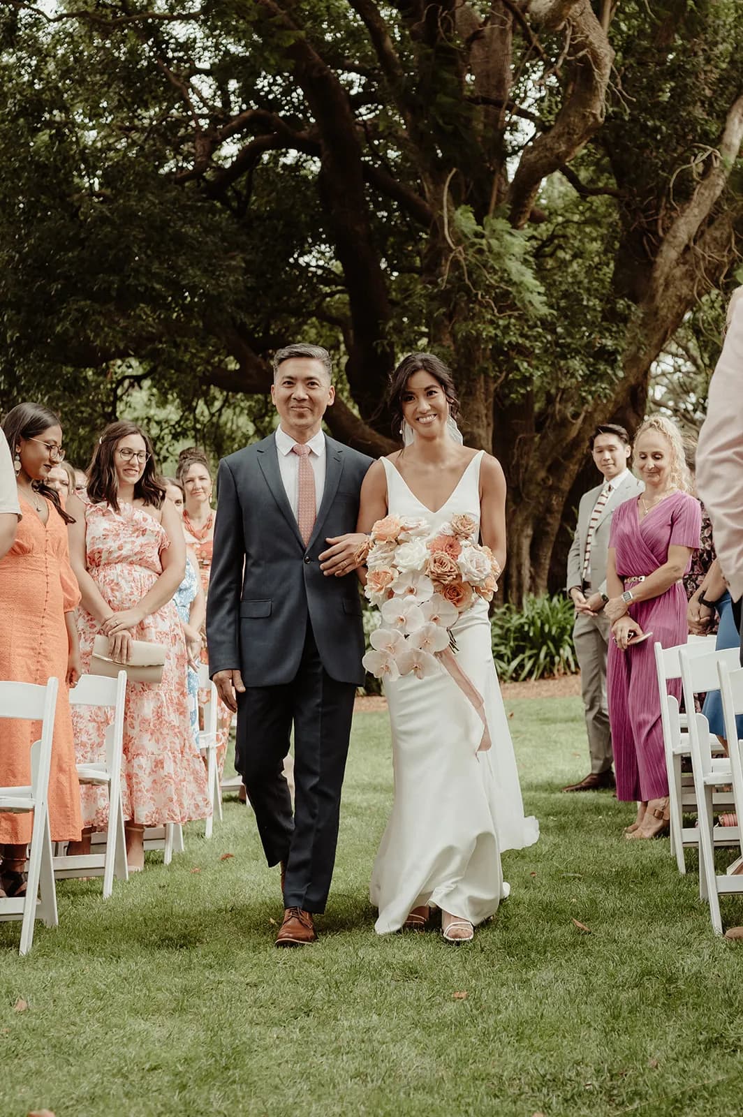 A bride, wearing a white gown and holding a bouquet of flowers, walks down an outdoor aisle arm-in-arm with an older man in a suit and tie. Guests, dressed in colorful attire, are seated on white chairs on either side, smiling and watching. Large trees frame the background.