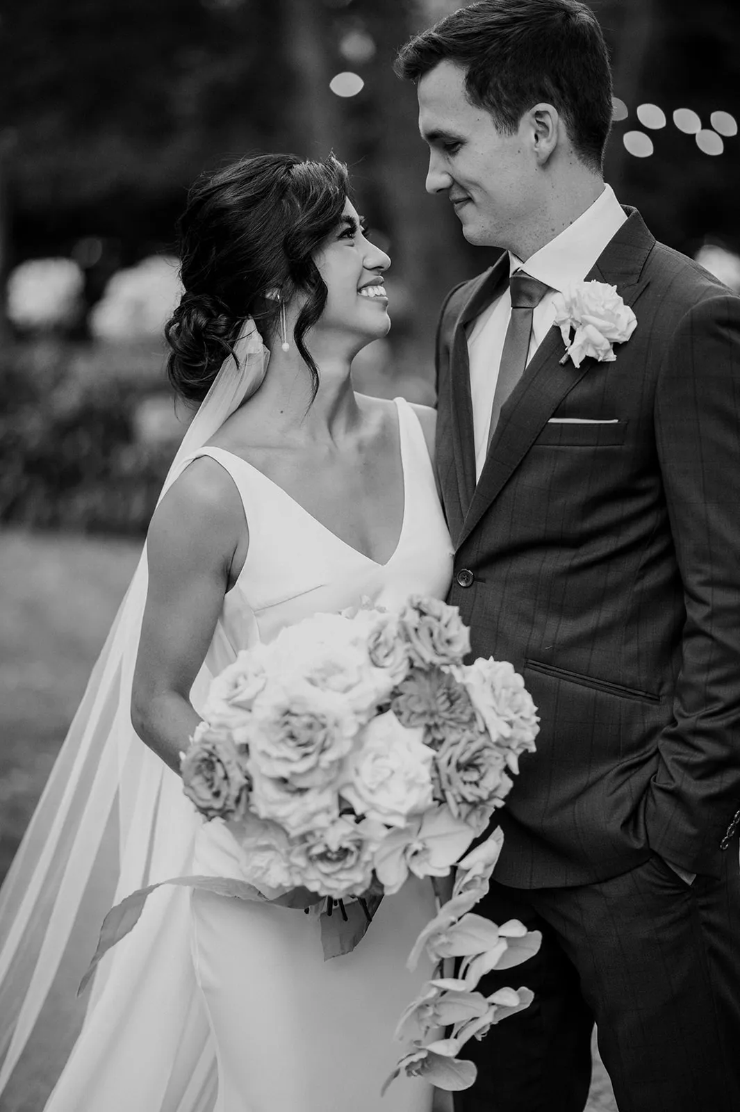 A newlywed couple stands close, smiling lovingly at each other. The bride, in a white dress and veil, holds a large bouquet of flowers. The groom wears a dark suit with a light-colored tie and boutonniere. The background is slightly blurred, suggesting an outdoor setting.