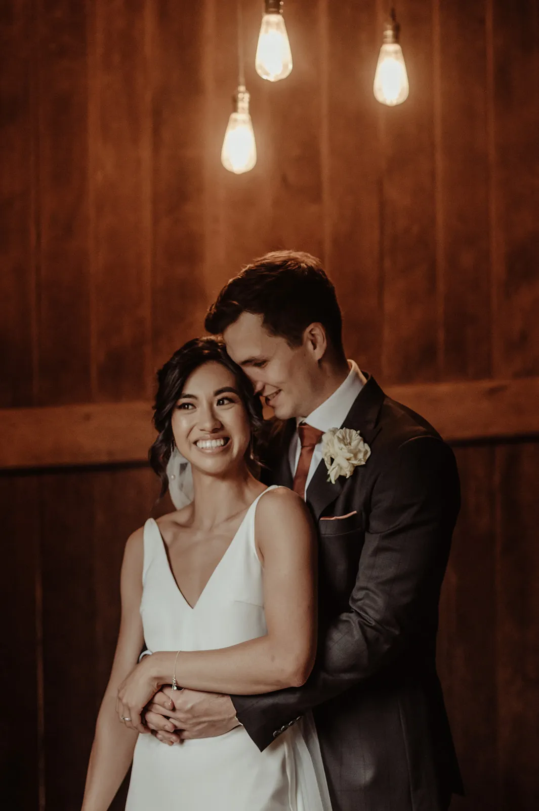 A couple is embracing in a warmly lit setting. The woman, wearing a white wedding dress, is smiling joyfully while the man, in a dark suit, stands behind her with his arms wrapped around her waist. Three hanging light bulbs illuminate the wooden background.