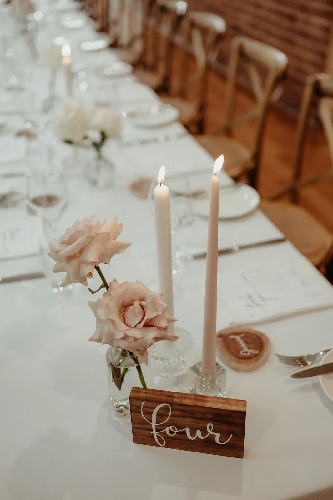 A long, elegantly set dining table with wooden chairs, featuring plates, silverware, and glasses. The centerpiece includes two lit white candles, two pink roses in small vases, and a wooden table number sign with the word "four" written on it.