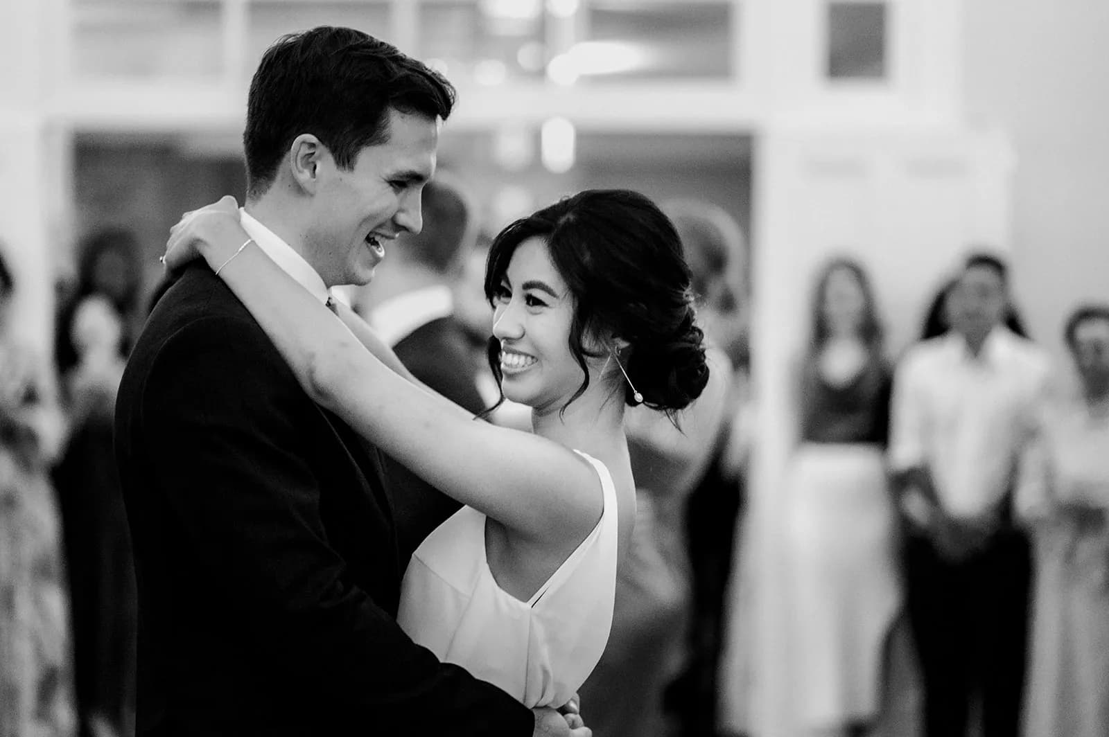A couple shares a joyful dance, smiling and holding each other closely in a black-and-white photograph. The woman is wearing a white dress, and the man is in a dark suit. A blurred crowd watches them in the background, adding to the celebratory atmosphere.