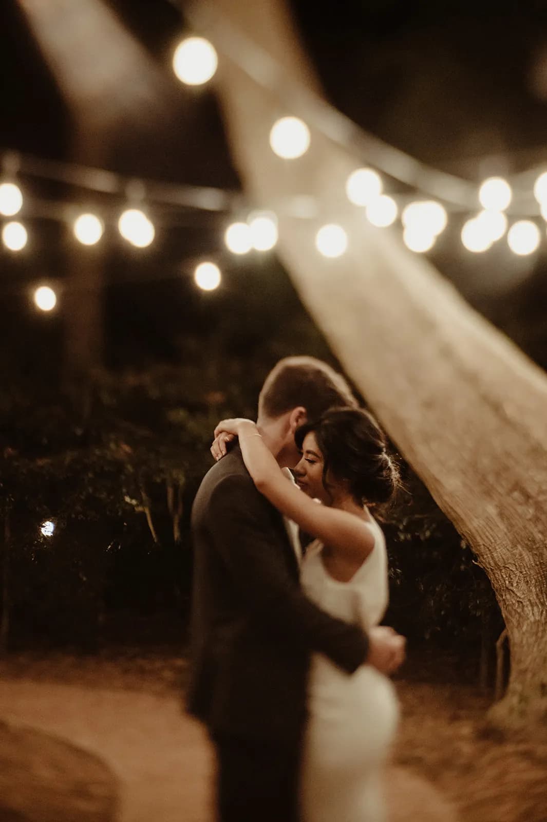 A couple embraces and dances under warm string lights in an outdoor nighttime setting. The background is blurred, emphasizing the intimate moment they share, illuminated softly by the glowing lights above. They appear to be alone together, creating a romantic atmosphere.