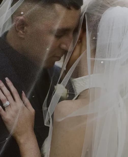A bride and groom share an intimate moment on their wedding day, nuzzling under a sheer veil. The bride's hand, adorned with a wedding ring, rests on the groom's chest. Both are dressed in formal wedding attire, and soft lighting creates a romantic ambiance.