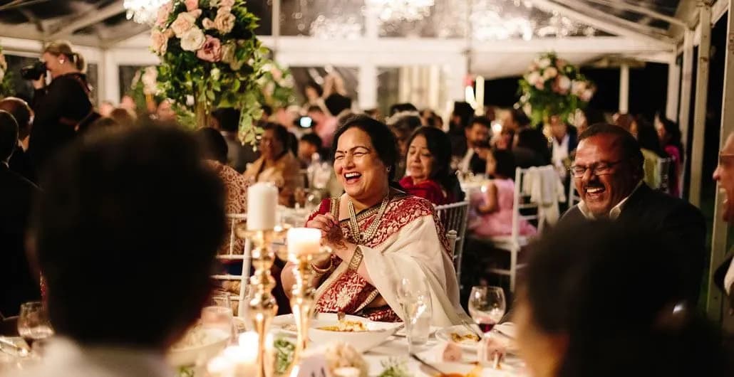 Guests seated around elegantly decorated tables, sharing laughter and joy at an event. A woman in a traditional outfit is clapping and smiling, surrounded by others equally engaged and smiling. The venue is adorned with floral arrangements and candles, creating a warm ambiance.