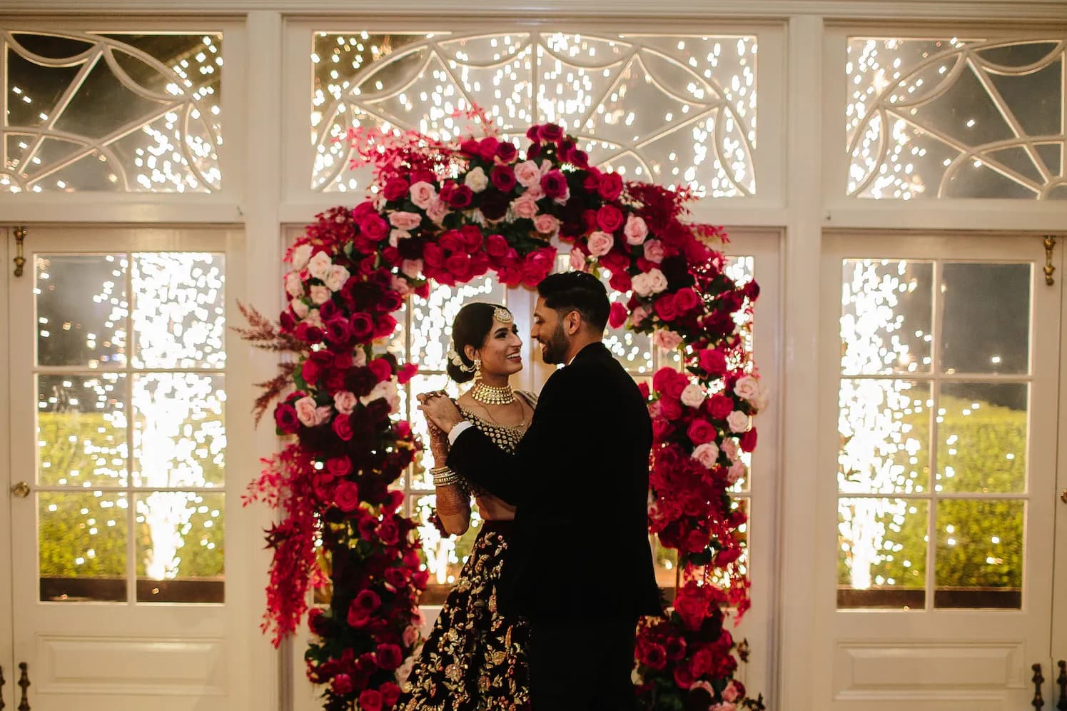 A couple dressed in formal attire stands embraced in front of a beautifully decorated floral arch with red and pink roses. The background features bright indoor fireworks, creating a celebratory and romantic atmosphere.