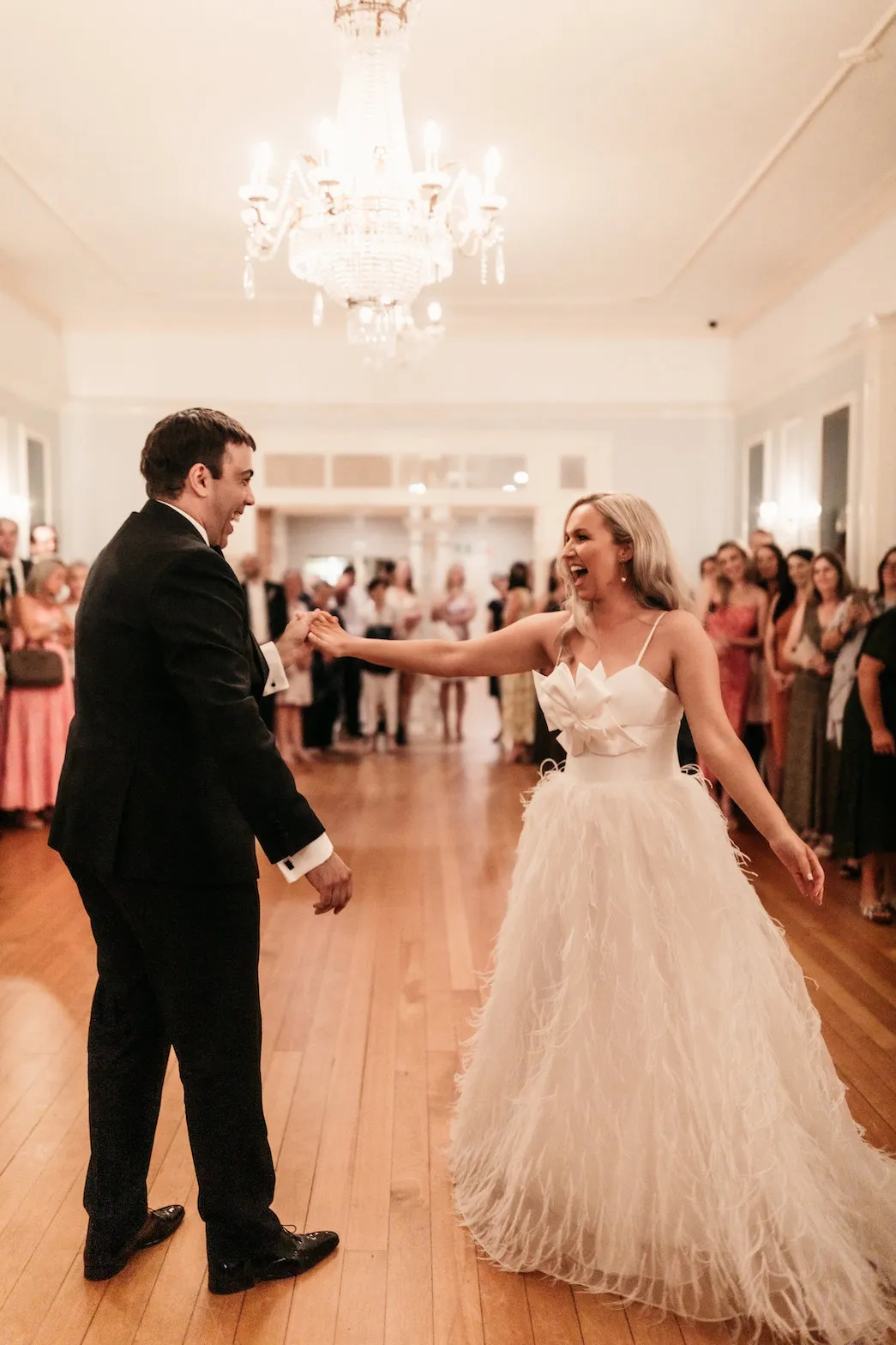 A newlywed couple dressed in elegant wedding attire share their first dance in a beautifully lit reception hall. The groom wears a black tuxedo, and the bride is in a white, textured gown. Guests stand smiling and watching in the background, under a sparkling chandelier.