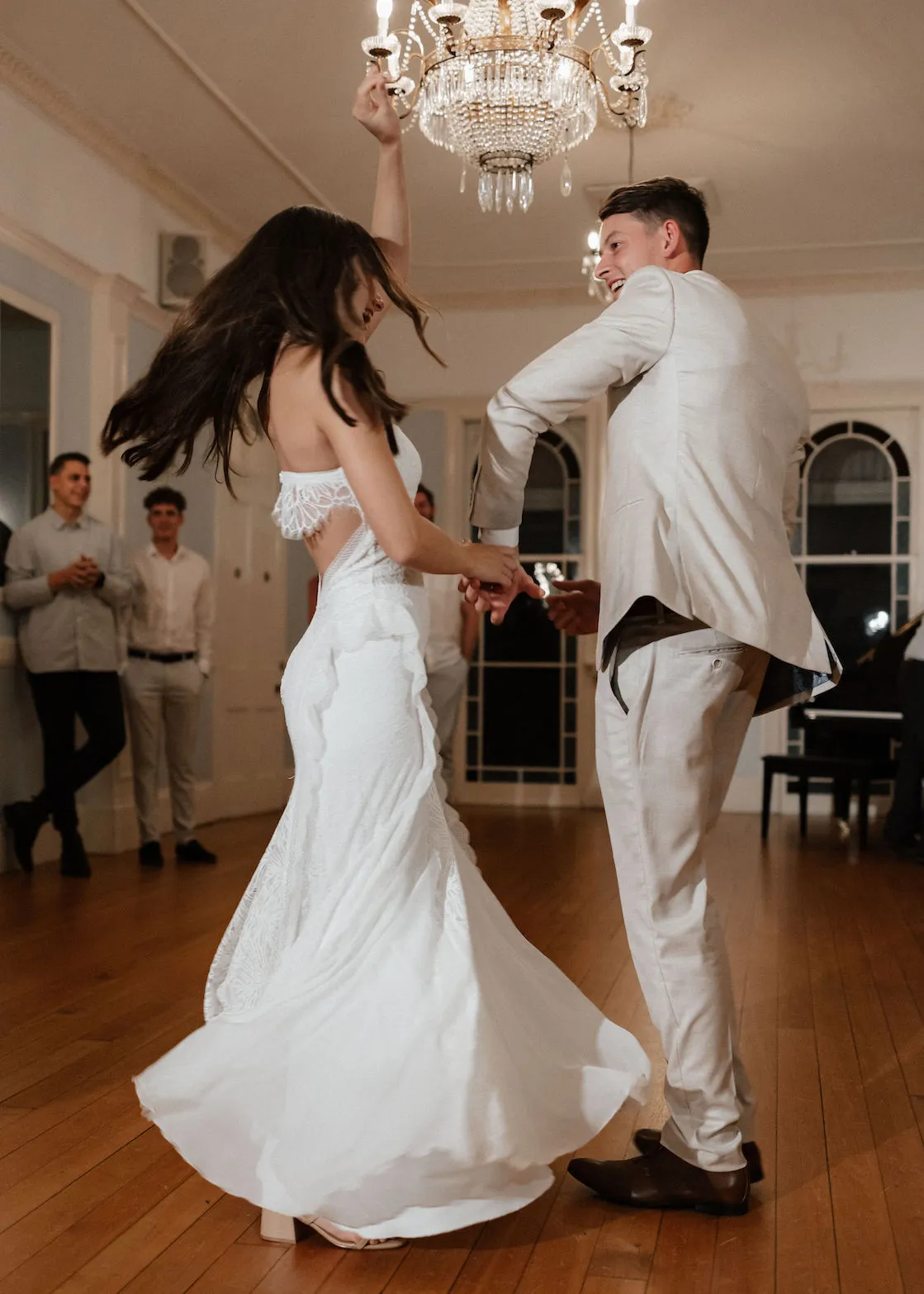 A bride in a white dress and groom in a light-colored suit dance under a chandelier in a spacious, elegant room with wooden floors. The bride’s dress twirls as they dance. Three guests in light-colored attire stand and watch in the background.