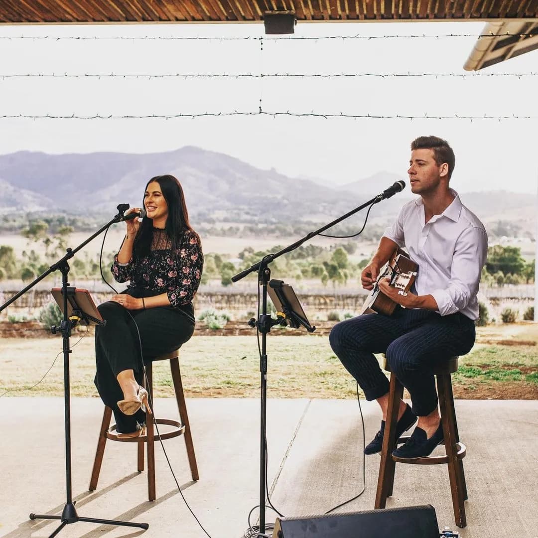 A woman sings into a microphone while sitting on a stool, and a man plays an acoustic guitar and sings beside her, also seated on a stool. They perform under a covered outdoor patio with a scenic countryside and mountains in the background.
