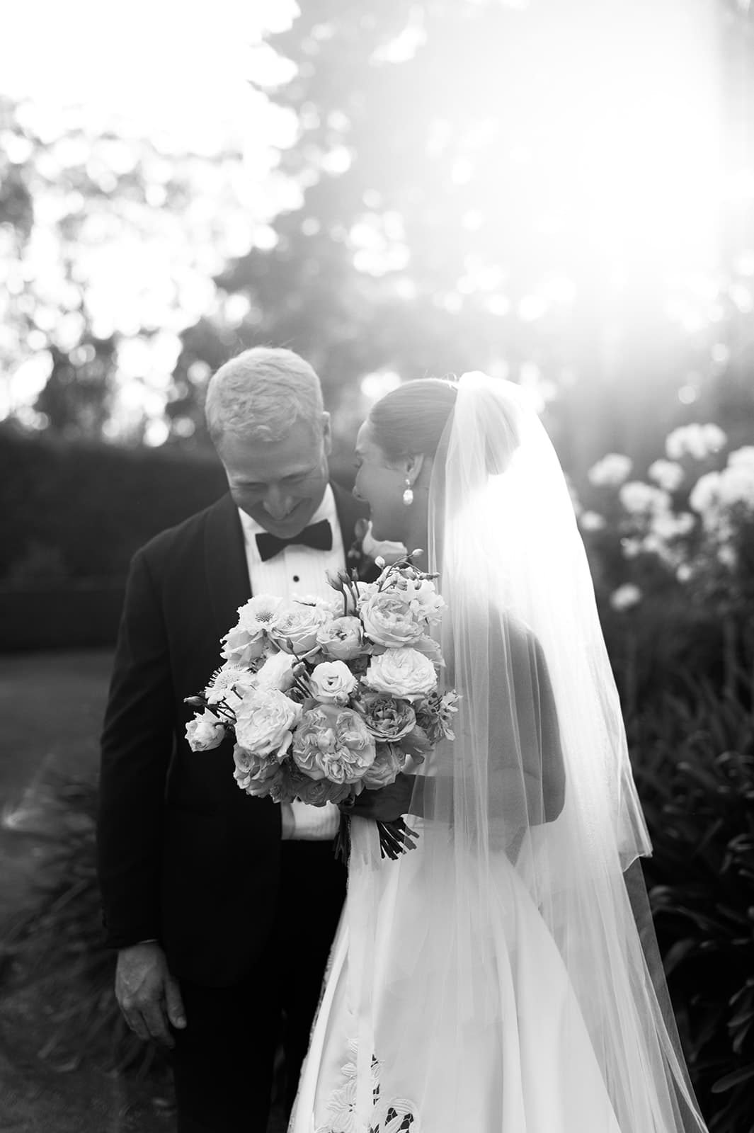 A black and white photo of a bride and groom on their wedding day. The bride, in a white gown and veil, holds a bouquet of flowers. The groom, in a black suit and bowtie, leans towards her. Sunlight illuminates them, creating a dreamy, romantic atmosphere.
