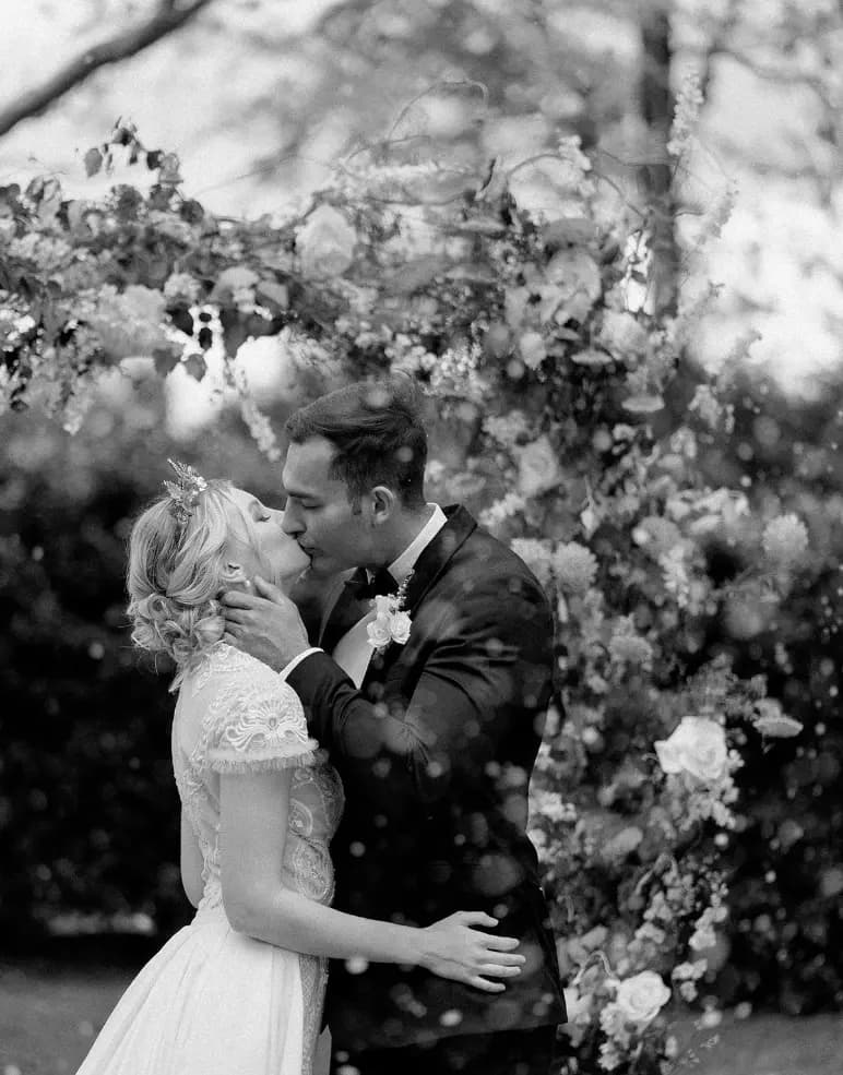 A black and white photo shows a couple sharing a romantic kiss outdoors on their wedding day. The bride, in a lacy dress and floral headpiece, and the groom, in a dark suit and bow tie, stand under a floral arch decorated with various blooms.