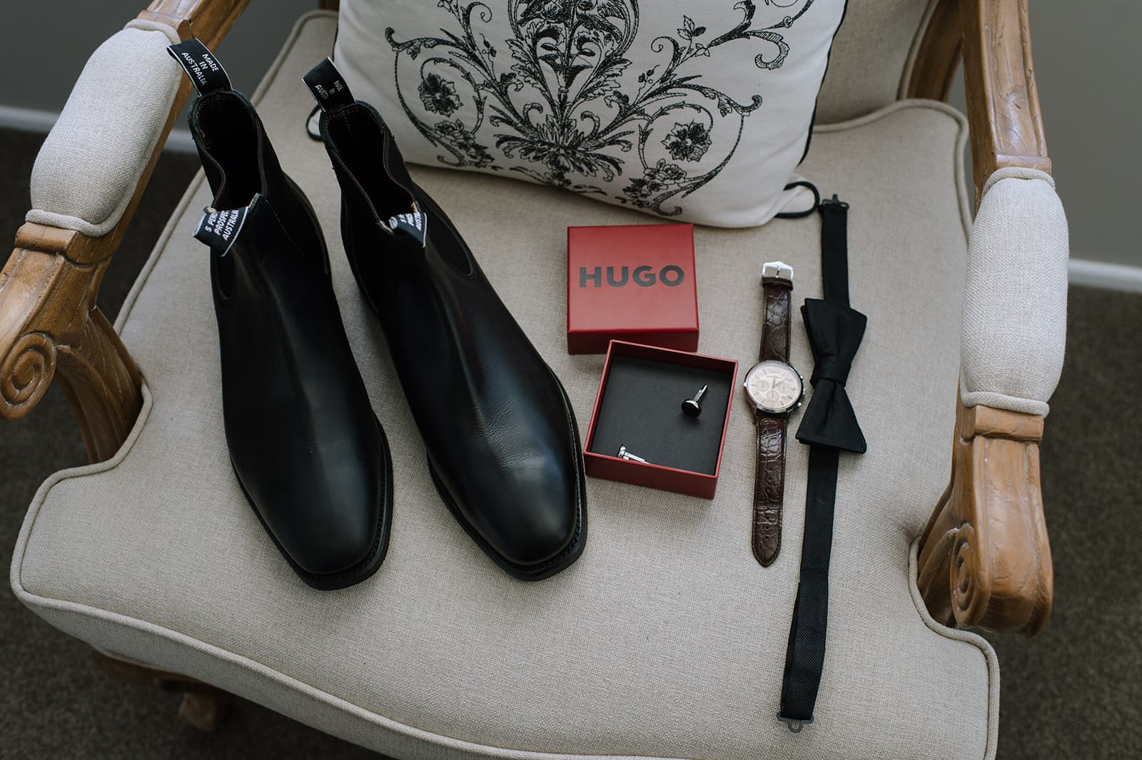 Black dress shoes, a red Hugo box with a ring, a brown leather strap watch, a black bow tie, and a white and black patterned cushion are neatly arranged on a beige cushioned wooden chair.