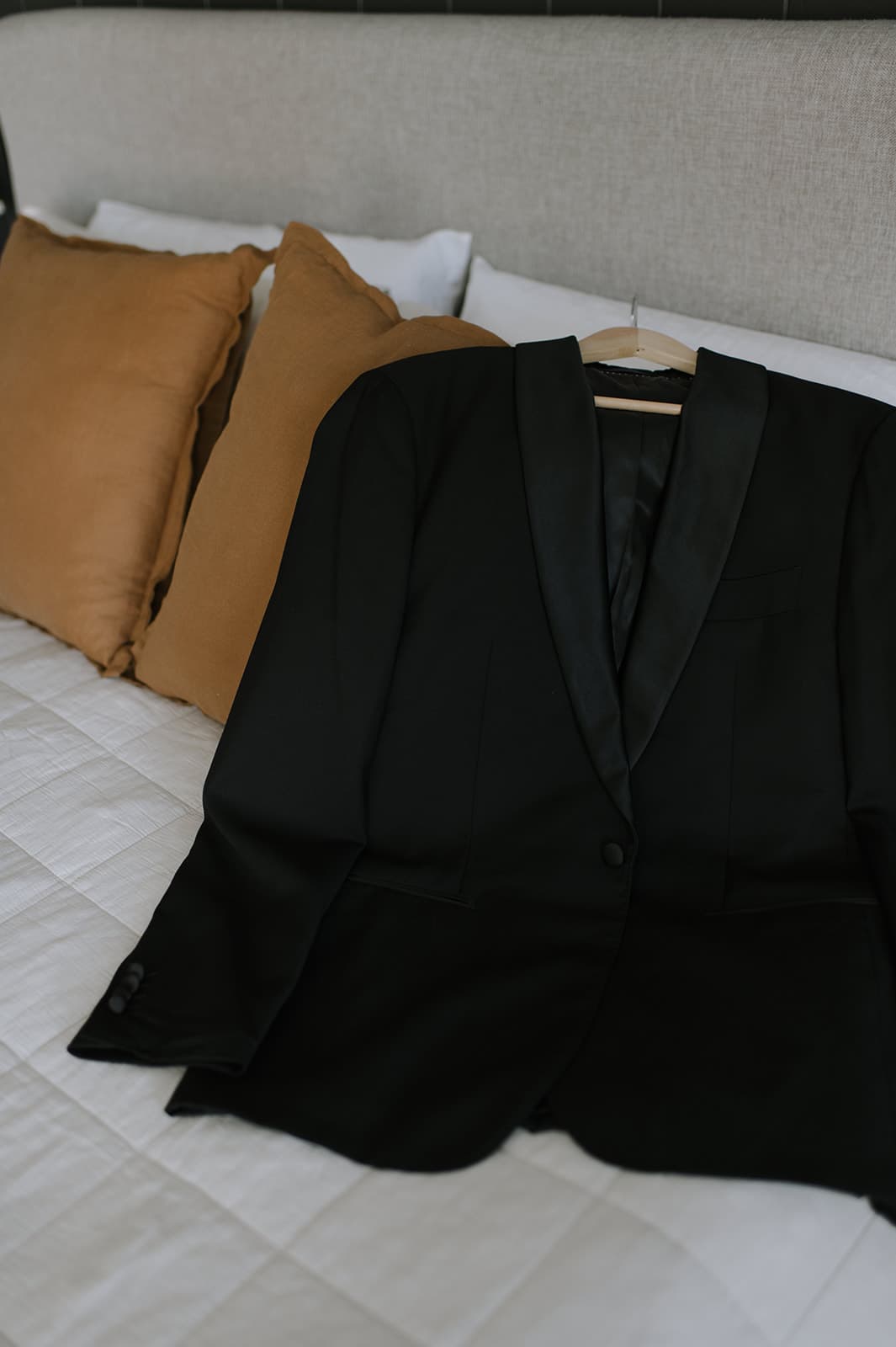 A black suit jacket is laid out on a white quilted bedspread with a beige headboard. Behind the jacket are three brown pillows neatly arranged against the headboard. The overall setting looks tidy and prepared.