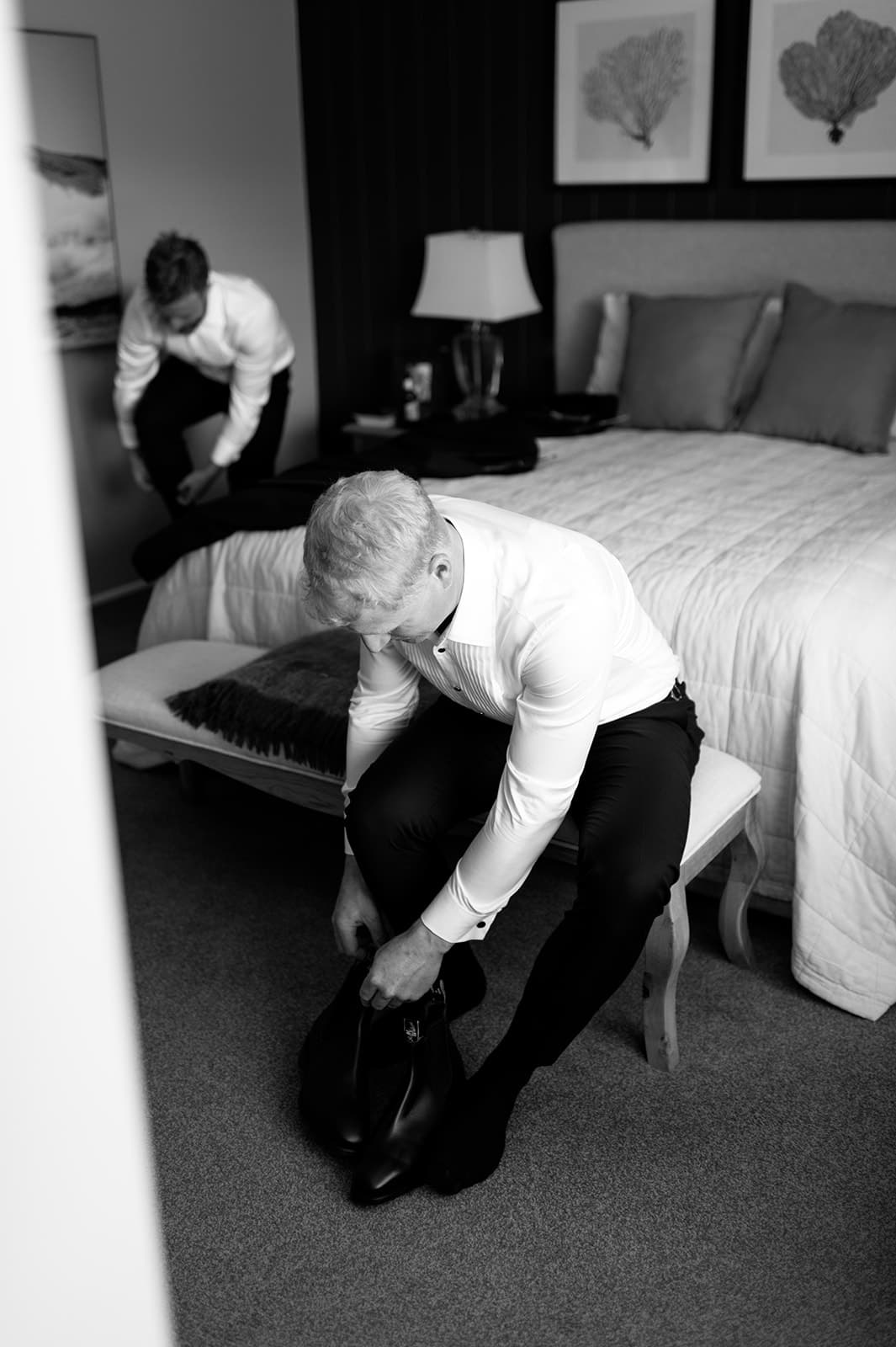 Black and white image of two men getting dressed in a bedroom. One man is seated on a bench near the bed, tying his shoe. The other man is in the background, fixing his pants. The room features a neatly made bed, a lamp, and framed artwork on the wall.