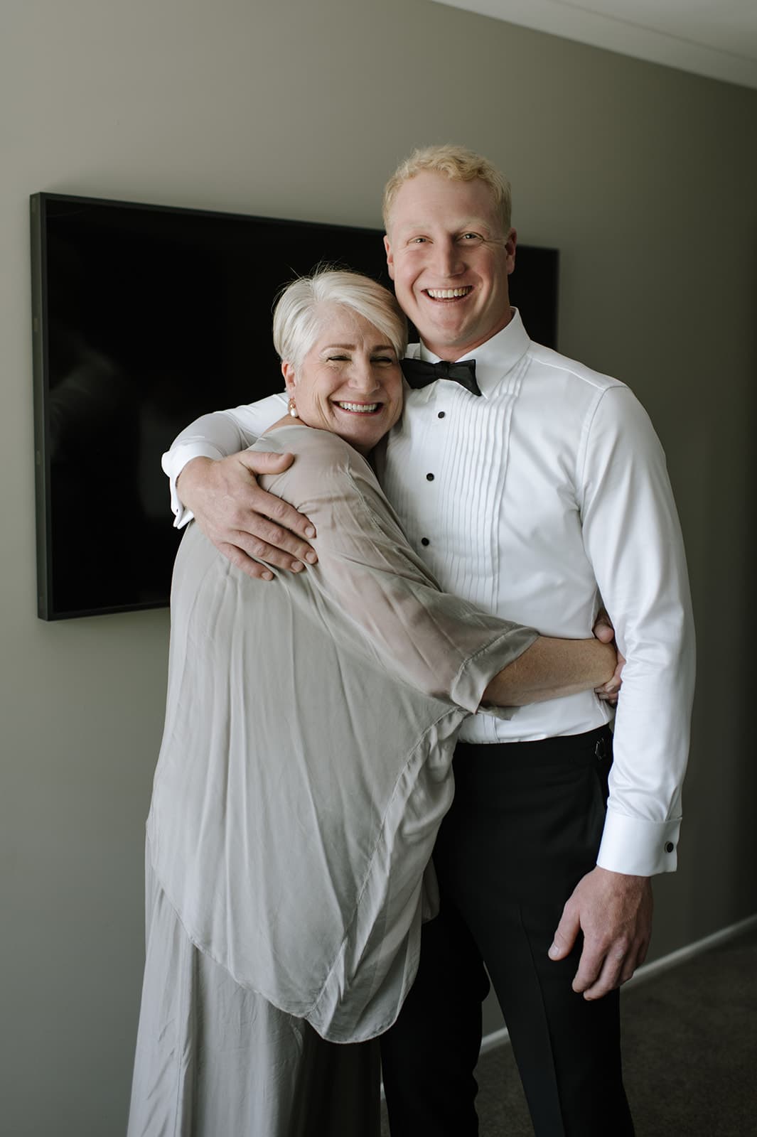 A smiling older woman with white hair embraces a younger man in a white dress shirt and black bow tie. They stand in front of a wall-mounted TV, both looking happy. The man holds her with one arm around her shoulder and the other hand resting on her upper back.